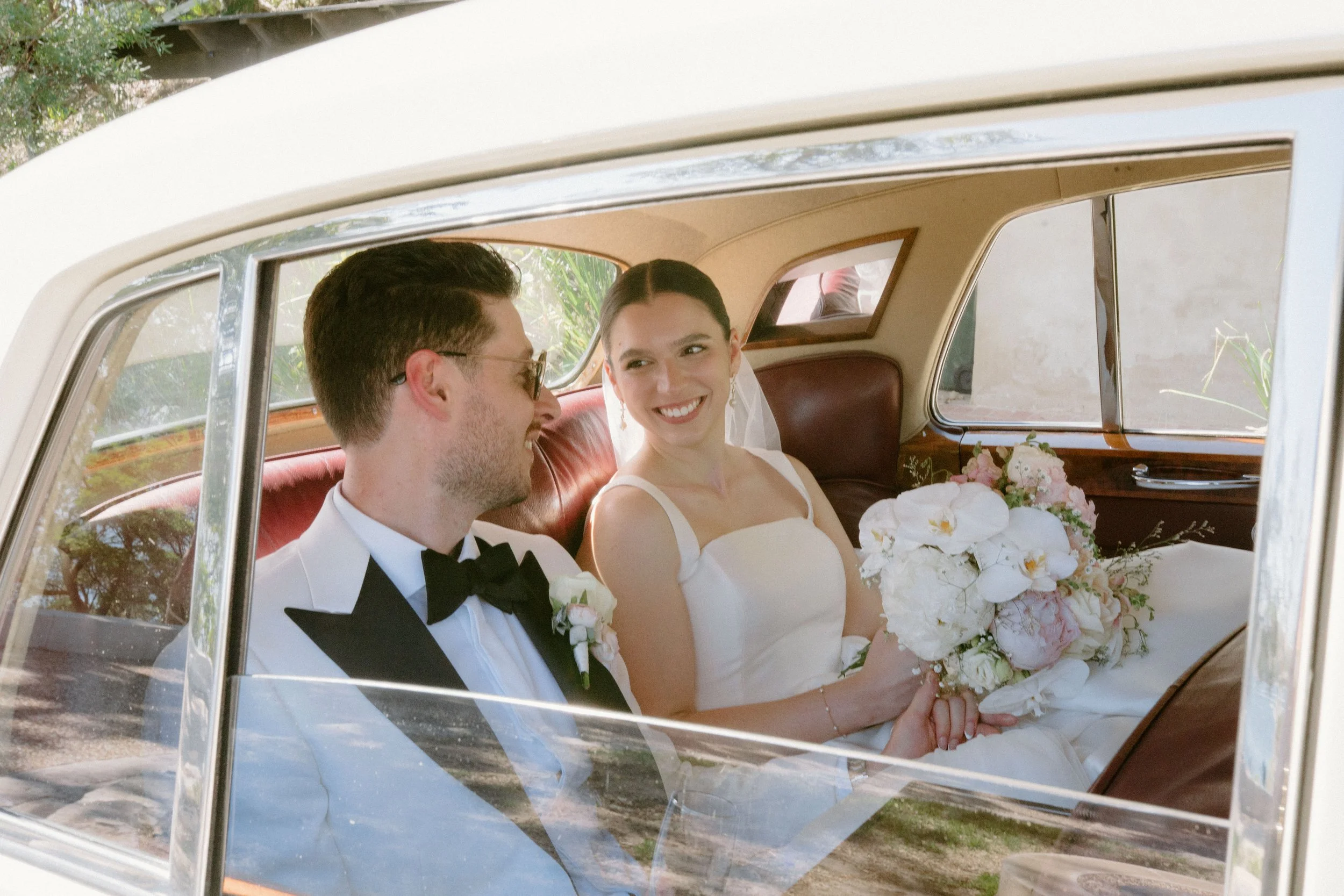 A bride and groom sitting inside a vintage car, smiling and holding a bouquet of flowers during their wedding.