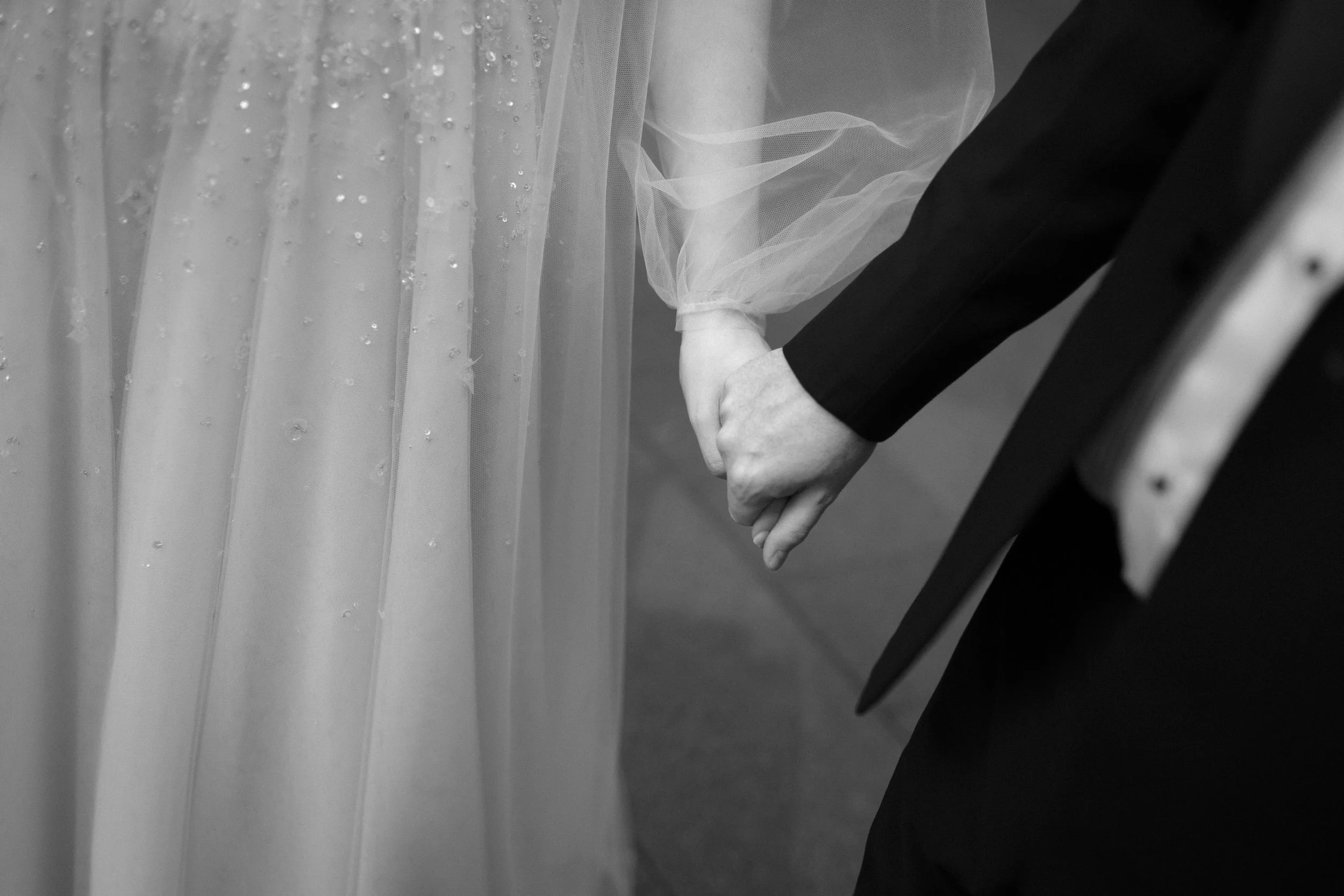 A black and white photograph of a bride and groom holding hands during a wedding ceremony. The bride is wearing a gown with a sheer overlay, and the groom is dressed in a dark suit.
