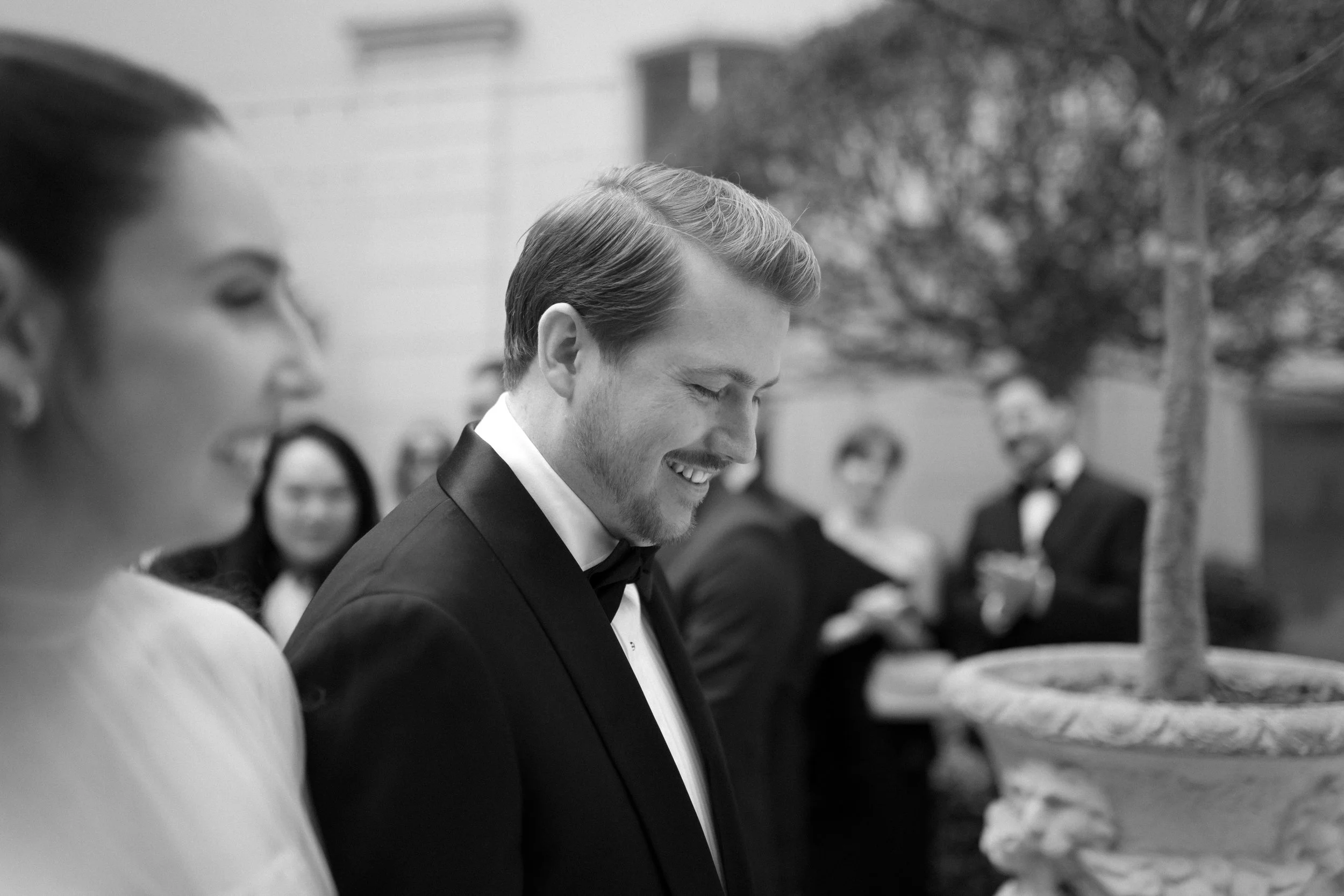 Black and white photo of a smiling man in a tuxedo at a formal outdoor event, with women and men in formal attire in the background.