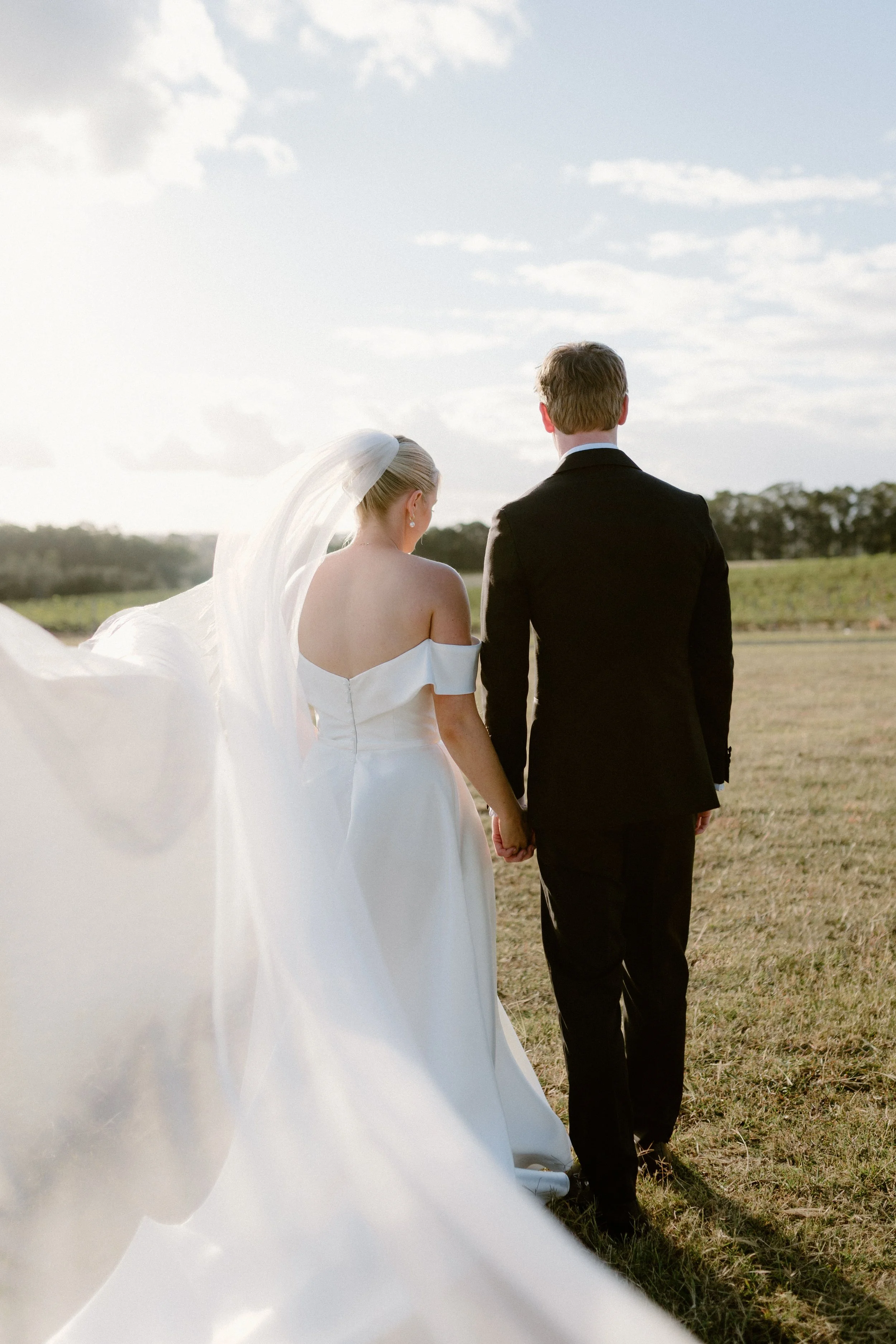 A bride and groom holding hands and walking away in an open field, with the bride's veil flowing in the wind, during sunset. Bimbadgen Palmers Lane Wedding, Hunter Valley