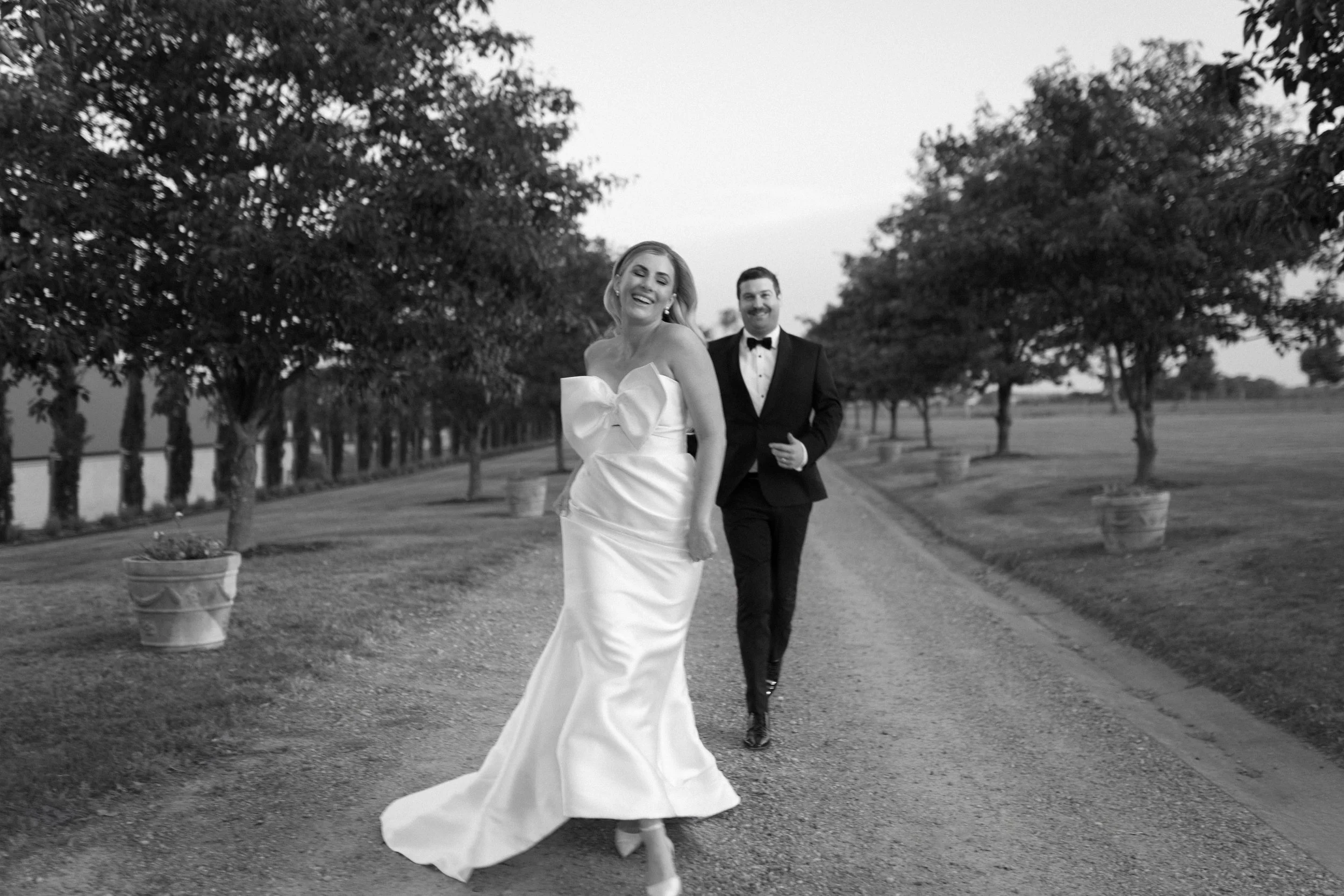 A black and white photo of a bride and groom walking down a path lined with trees and large potted plants. The bride is wearing a strapless wedding gown with a big bow on the front, and the groom is dressed in a tuxedo. Both are smiling and appear jo