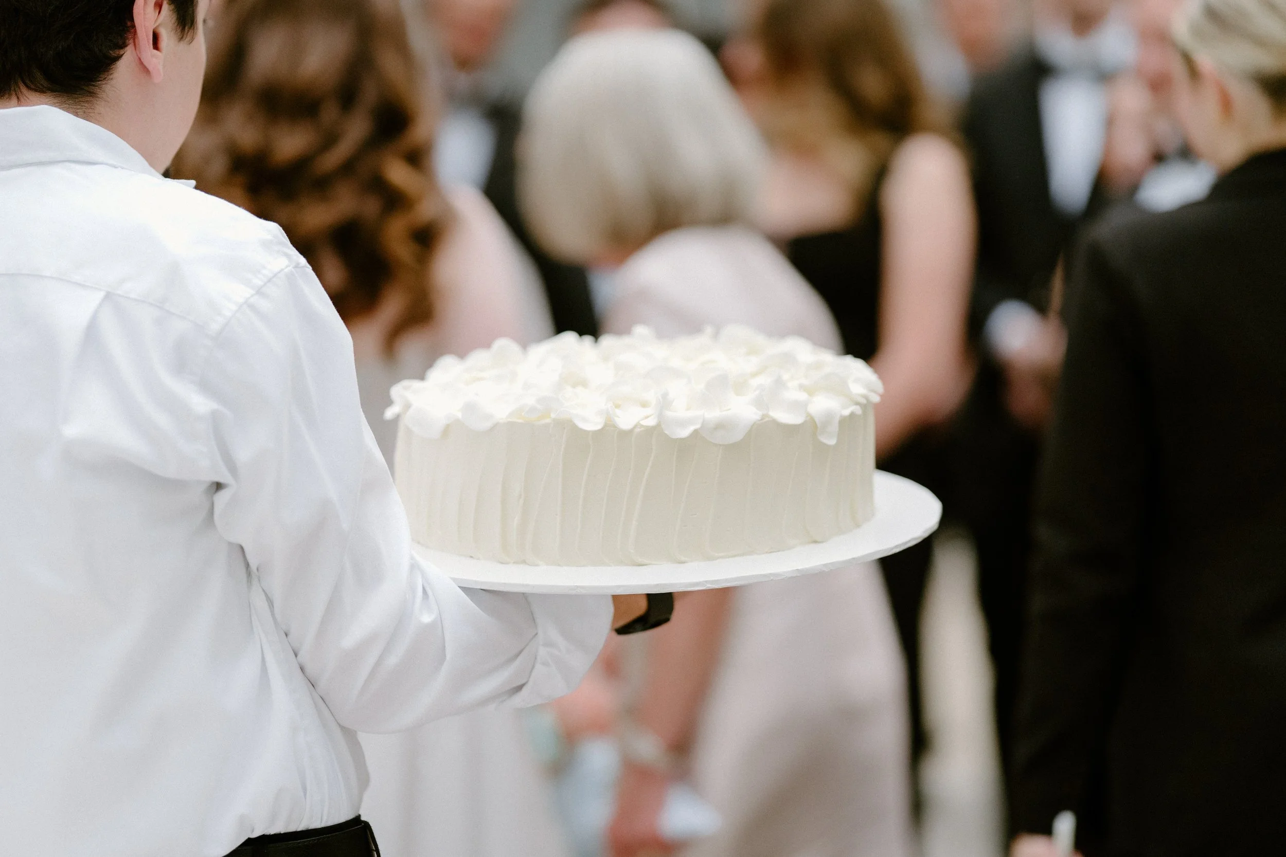 Person in white shirt holding a large white wedding cake with flower decorations, at a wedding reception with guests in formal attire in the background.