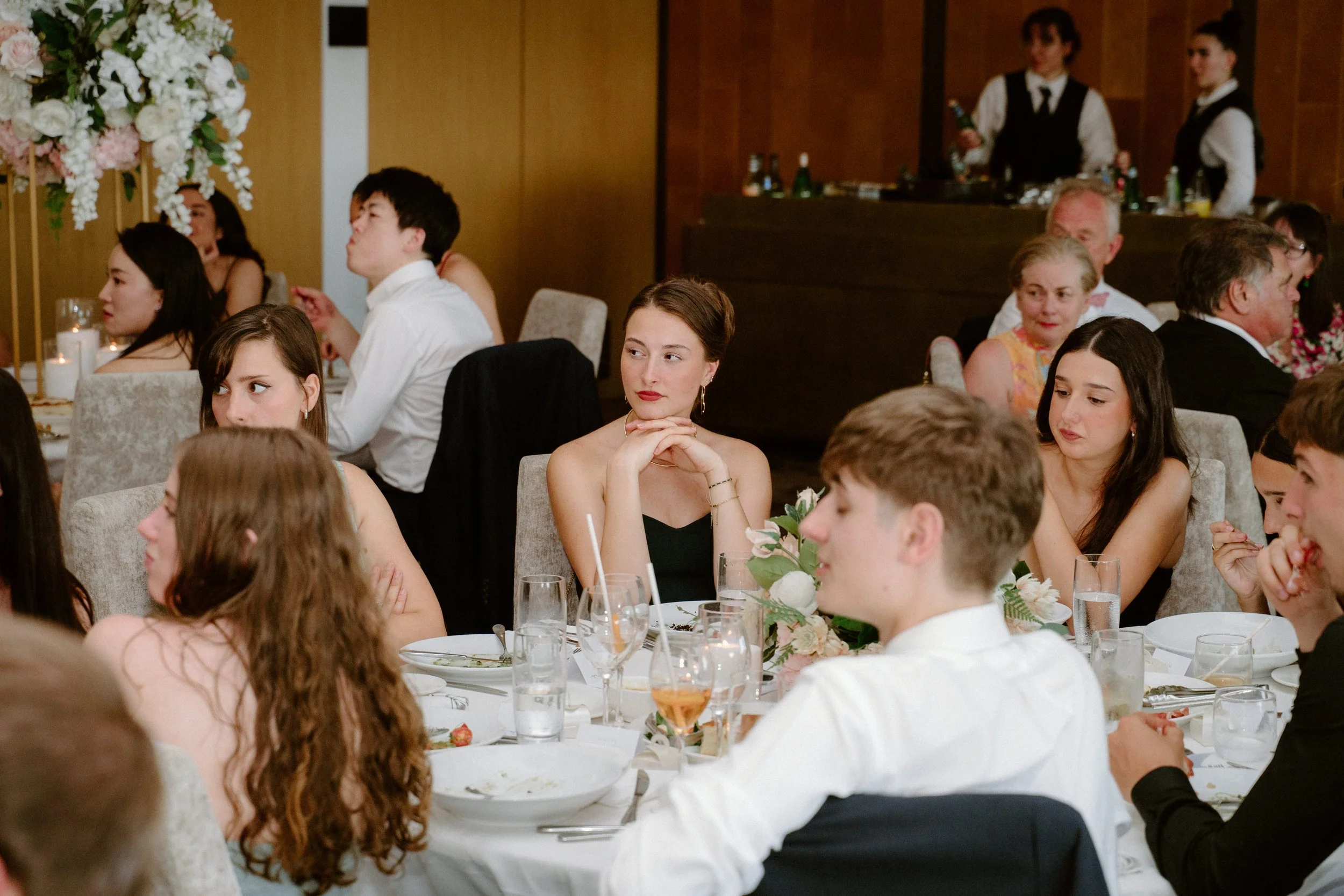 People seated at a formal event dining table, listening attentively; a woman in a black dress with jewelry rests her chin on her hands, surrounded by other guests and floral centerpieces.