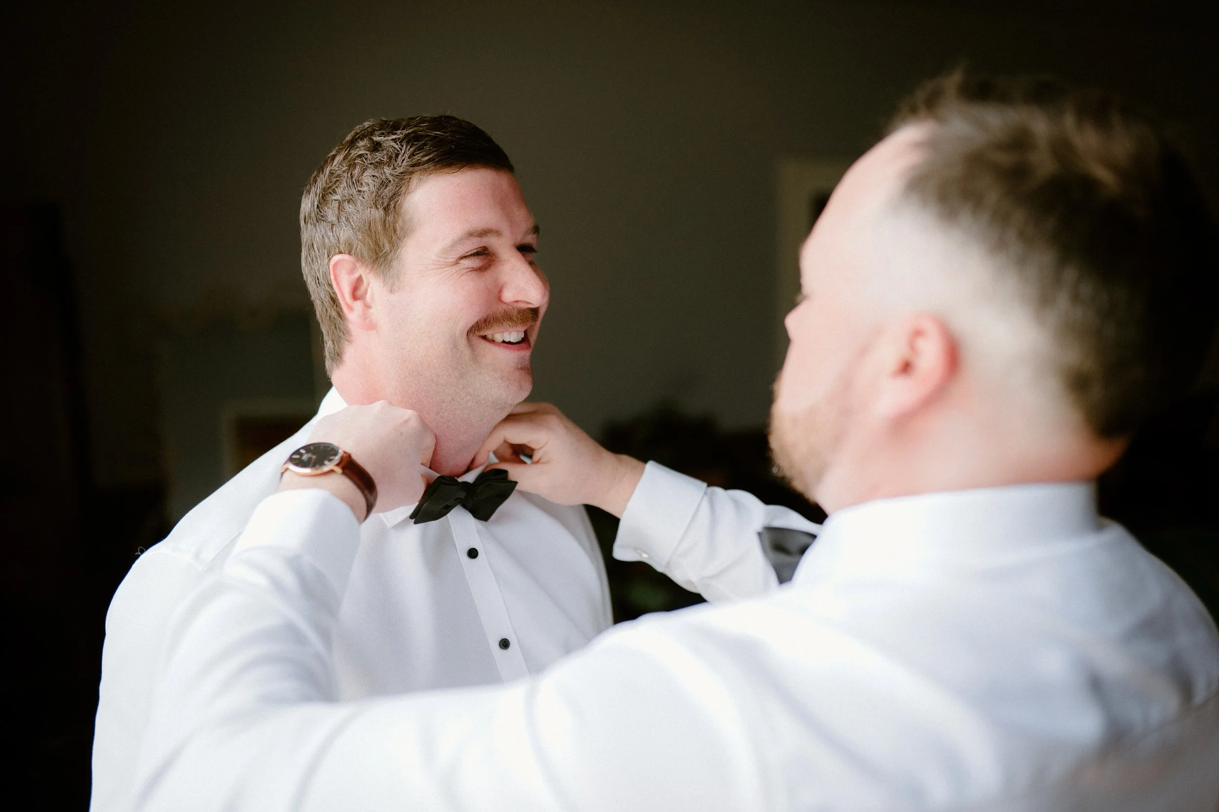 Two men in tuxedos smiling at each other, one adjusting the other's bow tie.