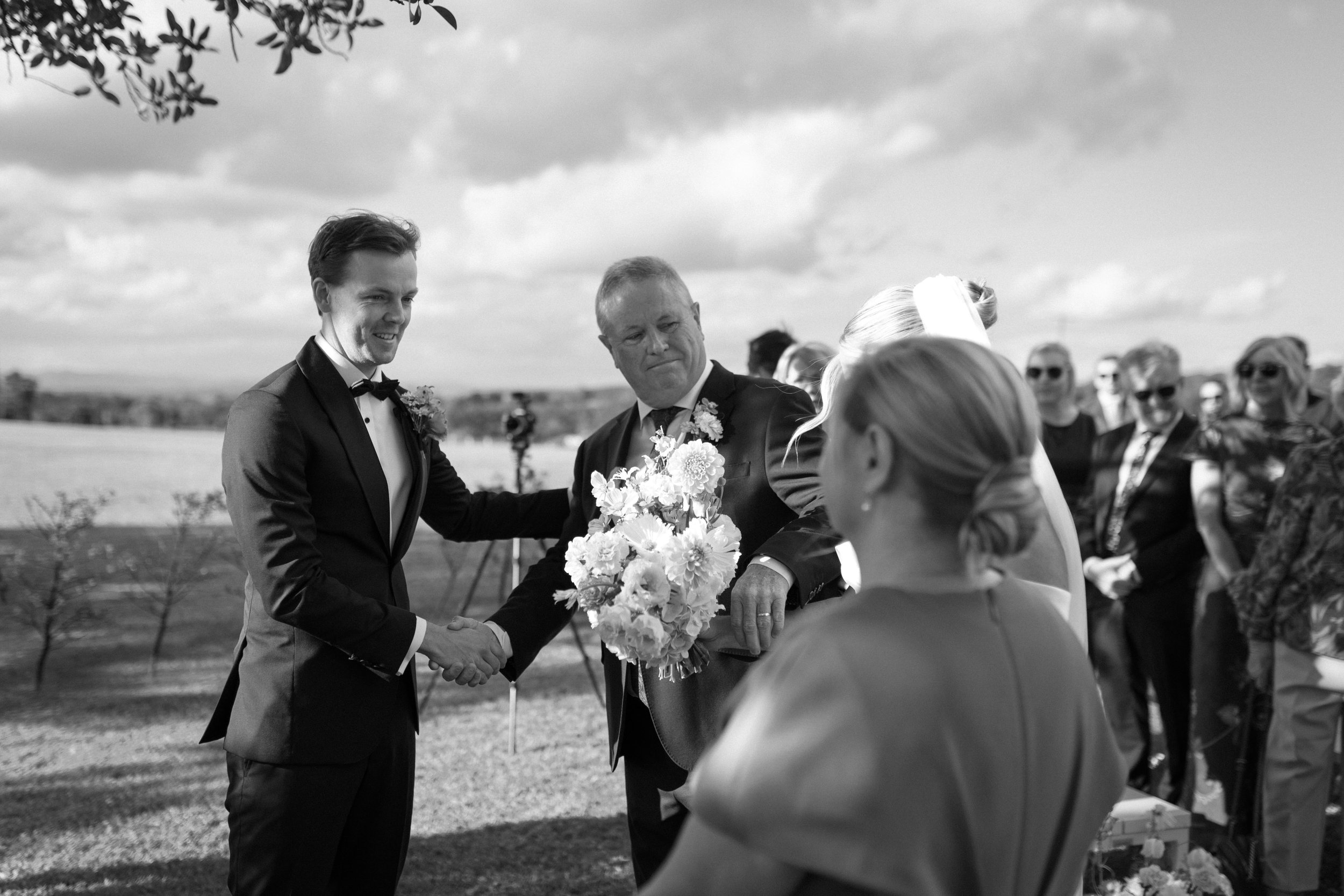 A black and white photo of a wedding ceremony outdoors. The groom, dressed in a tuxedo, is smiling and shaking hands with a woman who appears to be a bride, holding a bouquet of flowers. An officiant stands between them. There are guests in the backg