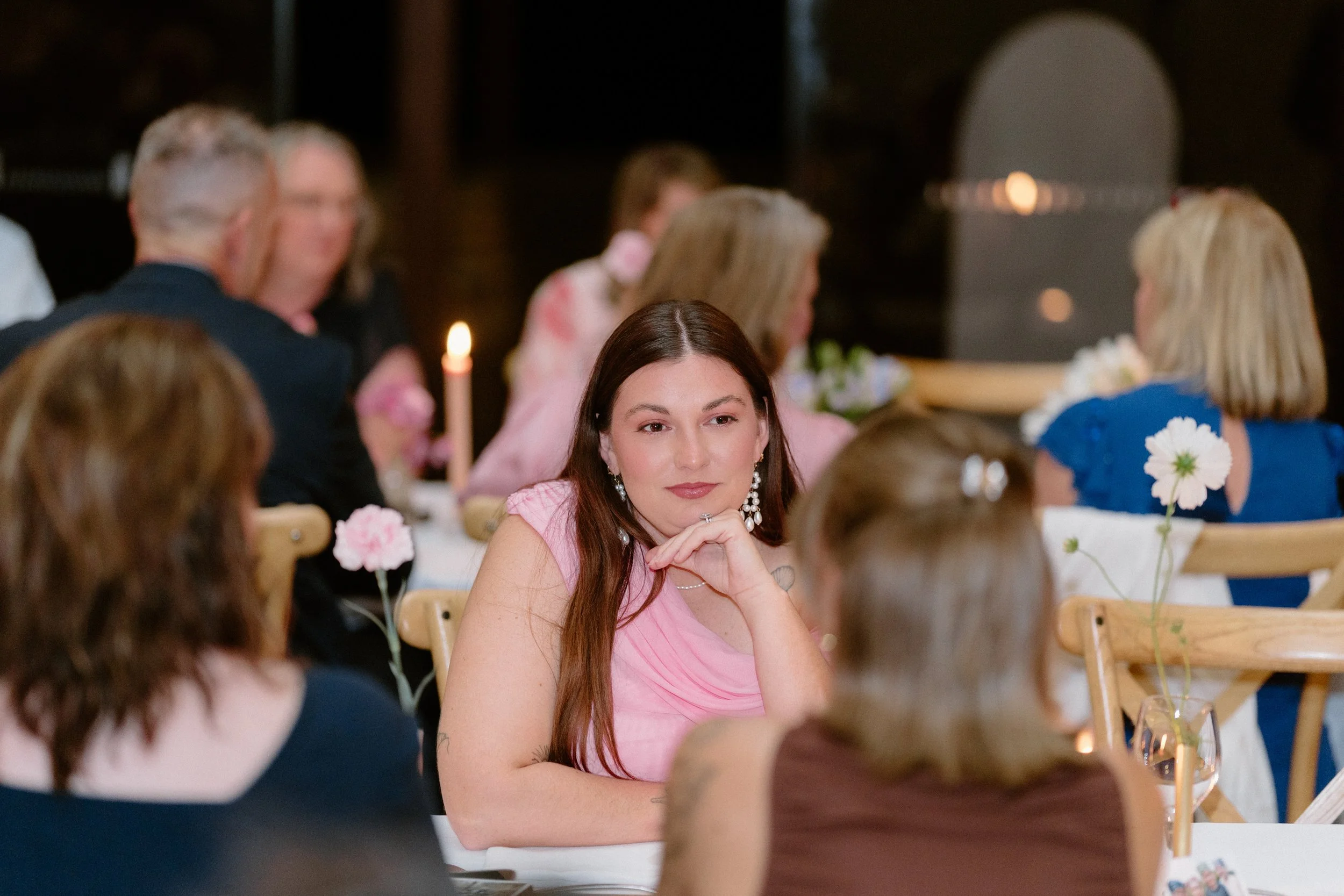 A group of people sitting at a table during a social gathering, with a woman in a pink top sitting in the center, resting her chin on her hand, surrounded by other guests, some of whom have flowers on their clothing.