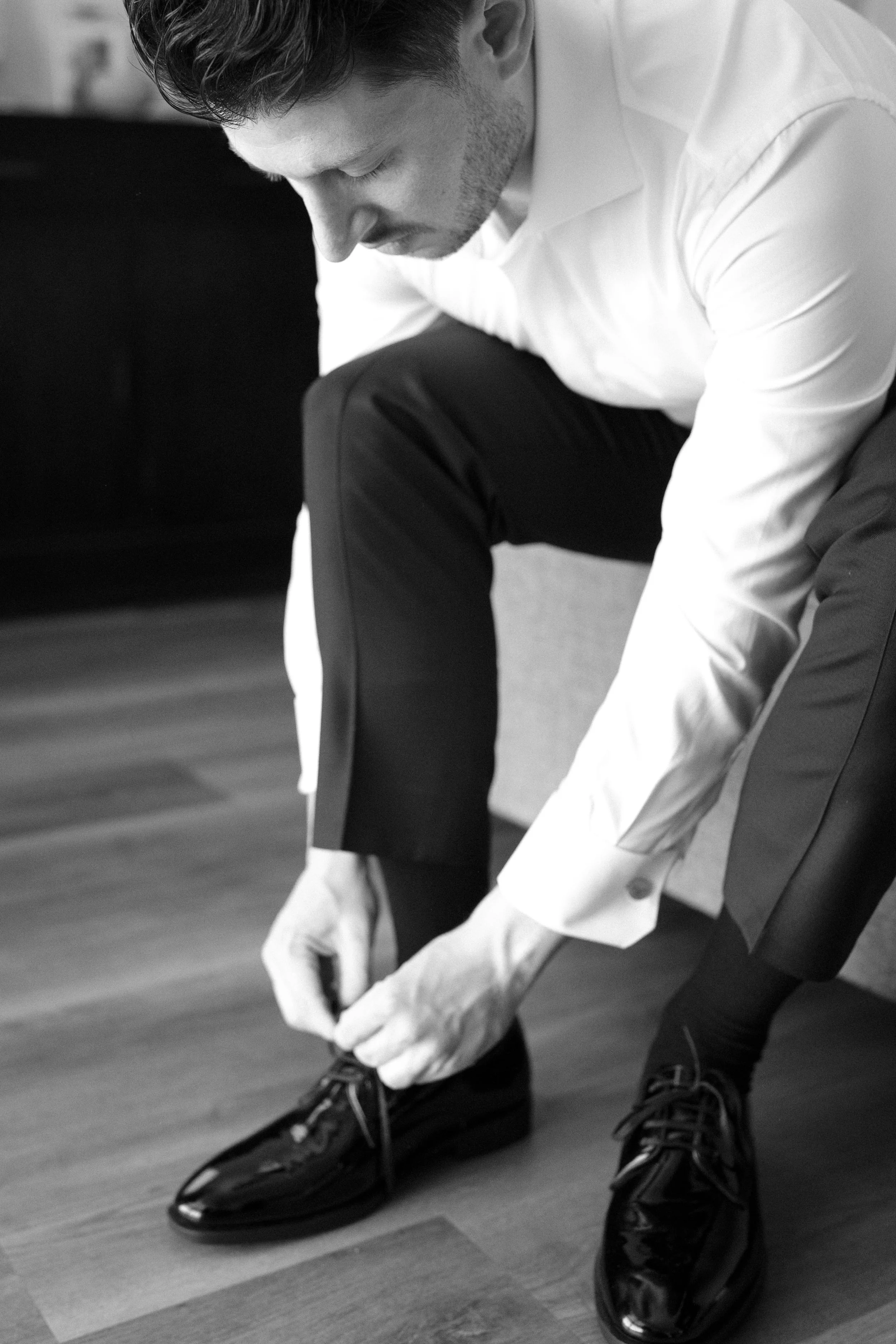 A man in formal attire is sitting on a couch, leaning forward, tying his black dress shoes.
