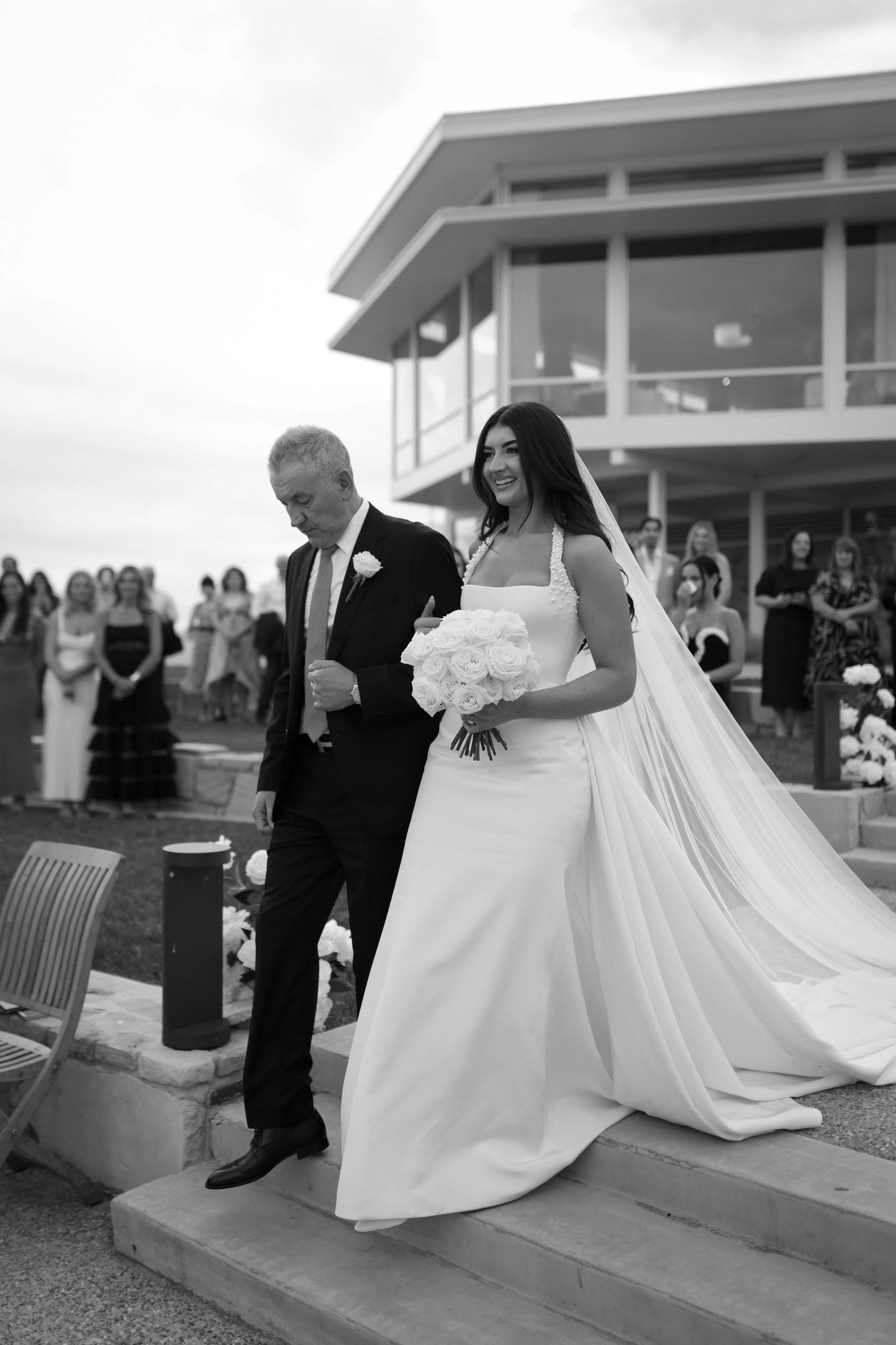 Black and white photo of a bride in a wedding dress holding a bouquet walking down steps with a man in a suit, with guests in the background, outside a modern building.