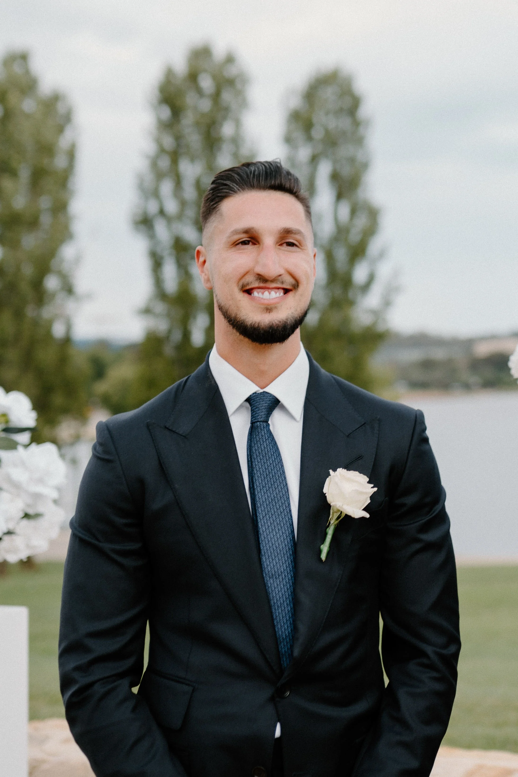 A man in a dark suit, white shirt, and blue tie, with a white flower boutonniere, smiling outdoors near a body of water and trees.