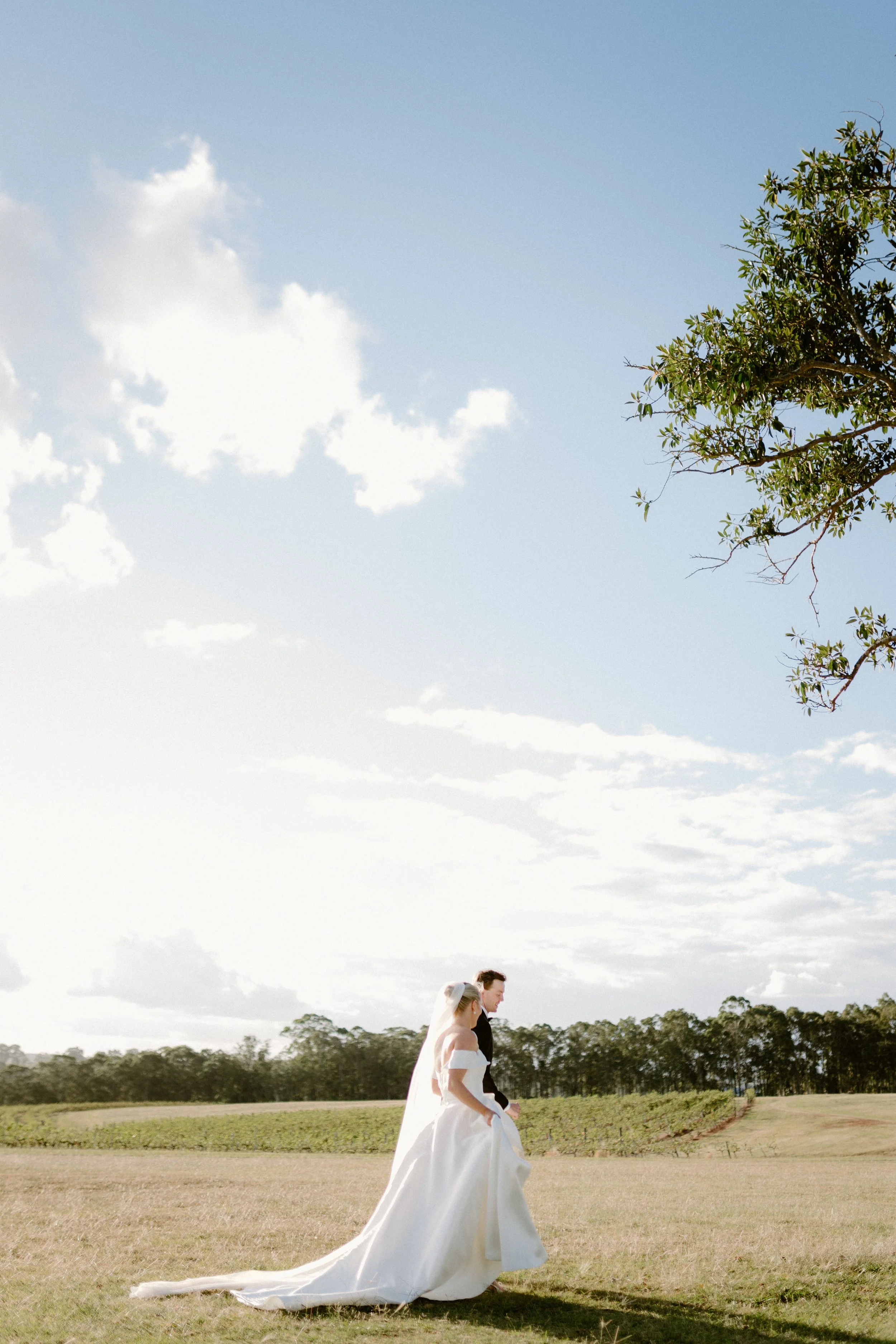 A bride and groom walking together in an open field, with a partly cloudy sky and trees in the background. Bimbadgen Palmers Lane Wedding, Hunter Valley