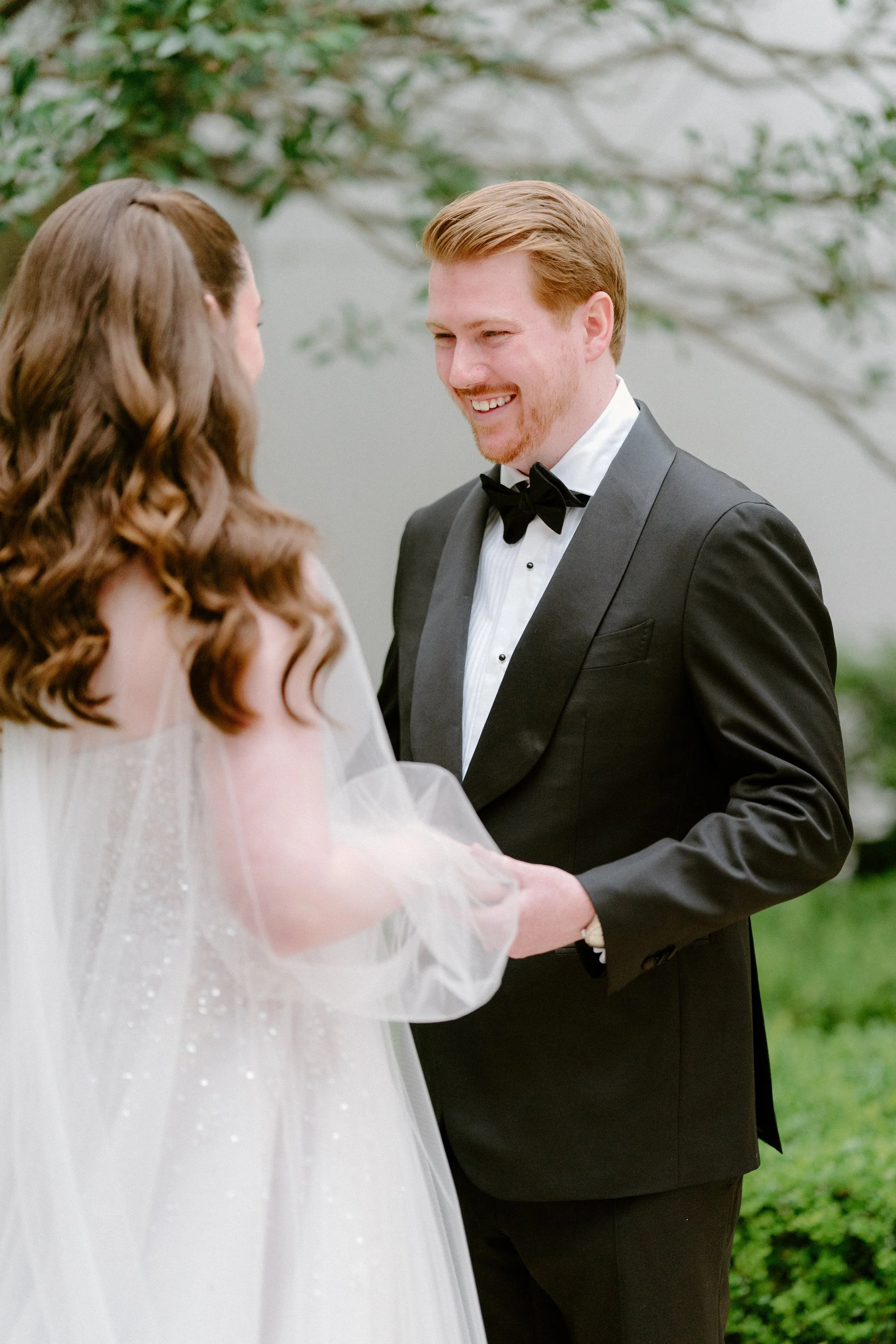 A groom in a black tuxedo holding hands with a bride in a white wedding dress during their outdoor wedding ceremony.
