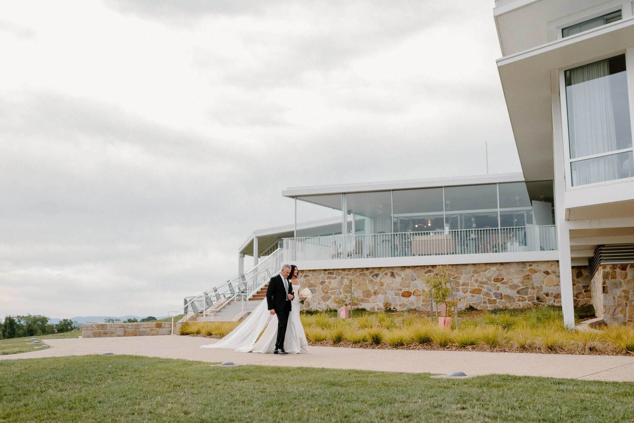 A bride and groom walking together outside a modern building with stone and glass exterior, on a cloudy day.