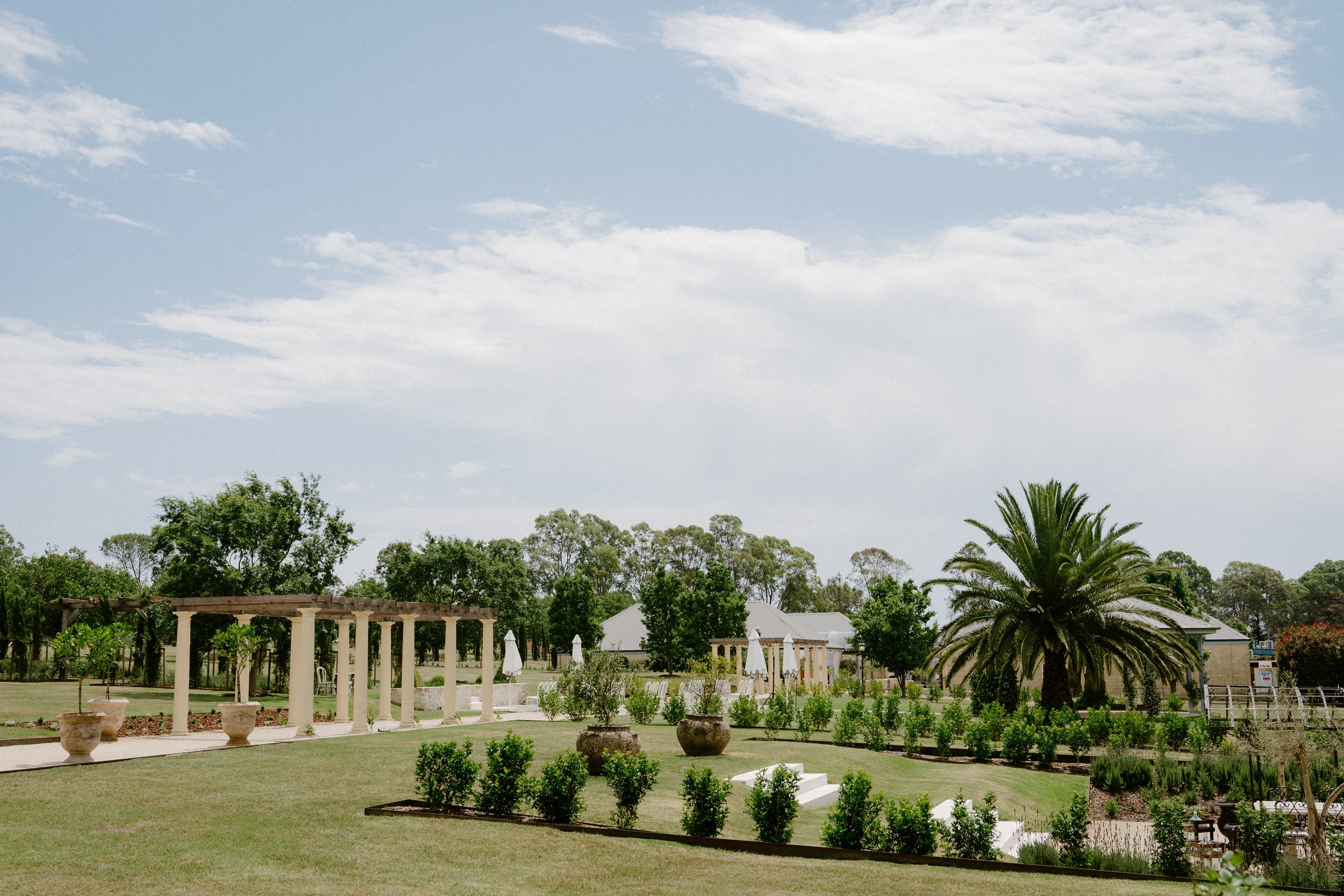 The Sienna, Hunter Valley Wedding Venue, Wedding Photos. A landscaped garden with potted plants, a large palm tree, and a pergola, under a partly cloudy sky.