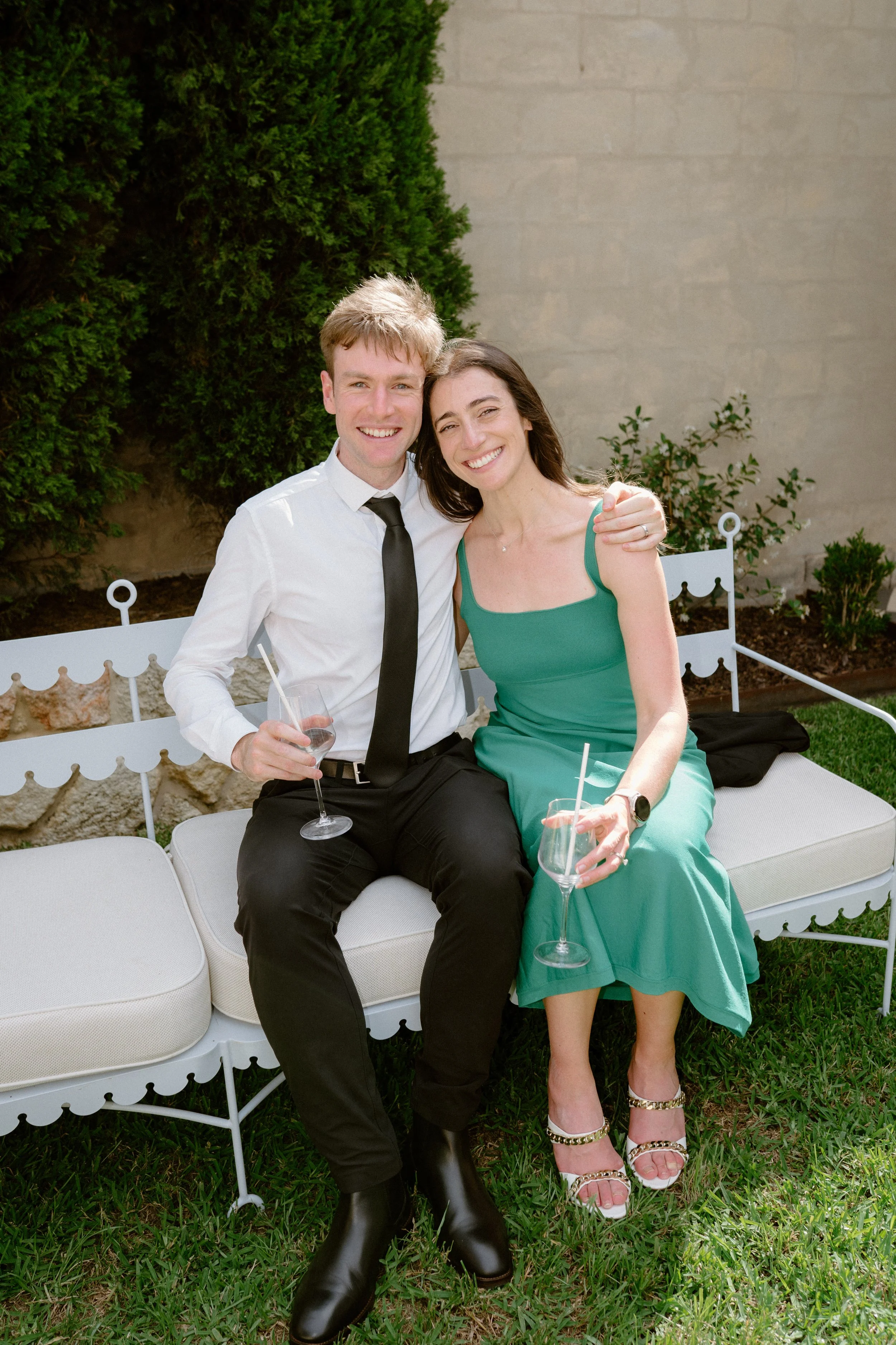 A smiling couple sitting on an outdoor white bench, holding glasses of wine, with a green bush and beige wall in the background.