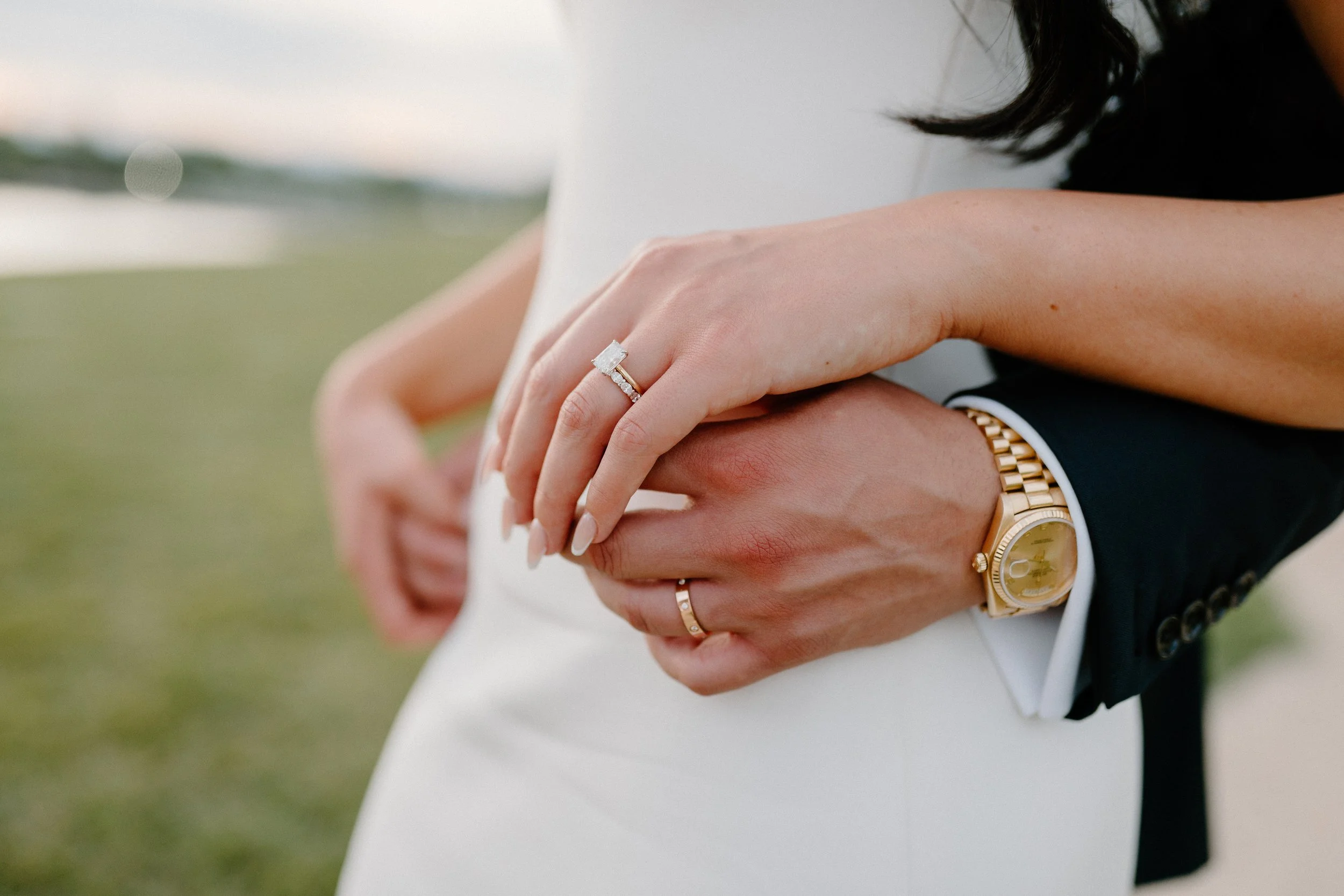A close-up of a bride and groom's hands intertwined. The bride wears an engagement ring, and the groom wears a gold watch and ring. The bride's wedding dress is white, and the groom's suit sleeve is visible.