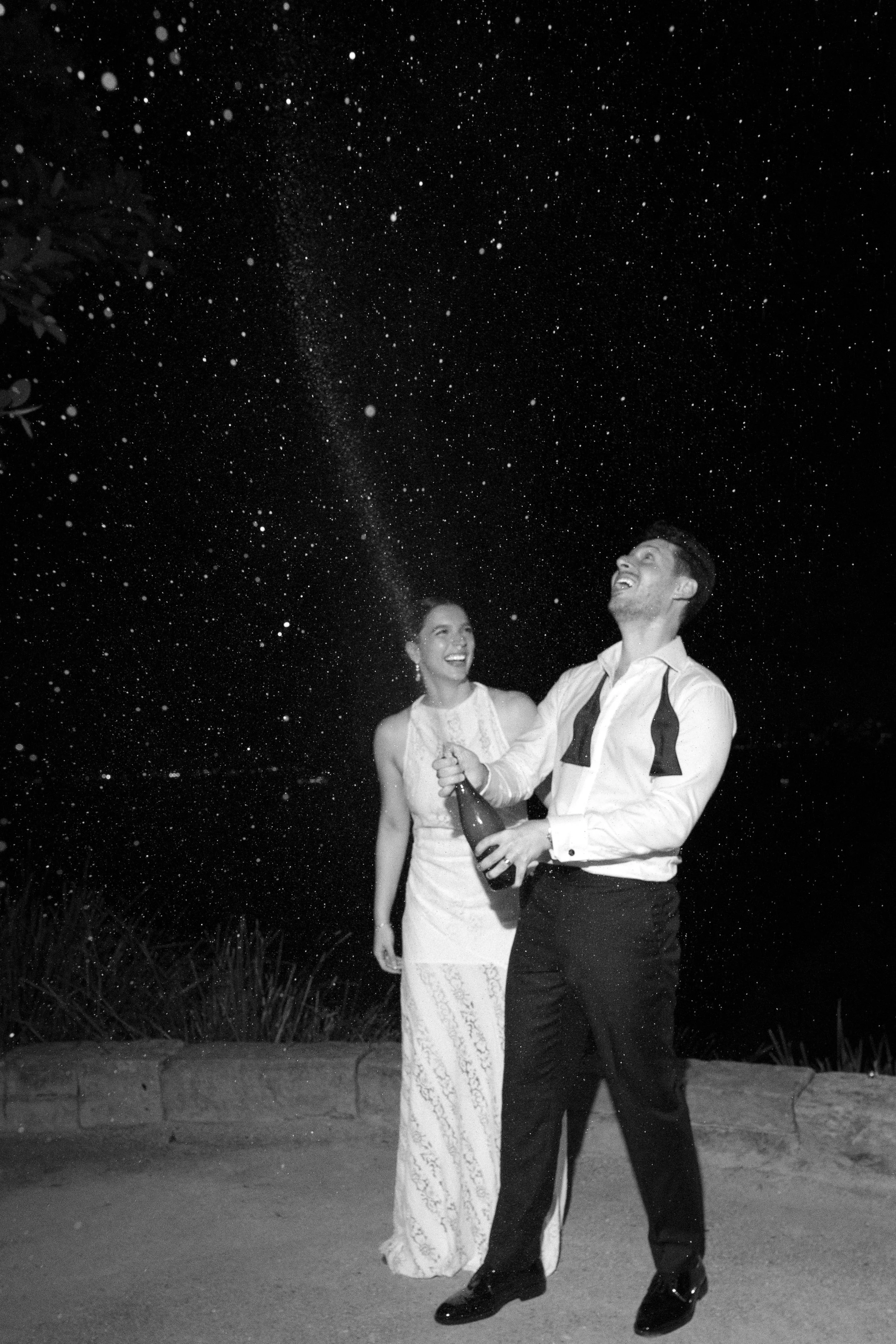 A black-and-white photo of a couple celebrating outdoors at night. The man is holding a champagne bottle, spraying champagne into the air, and the woman is laughing, wearing a long dress. The night sky with stars is visible above them.