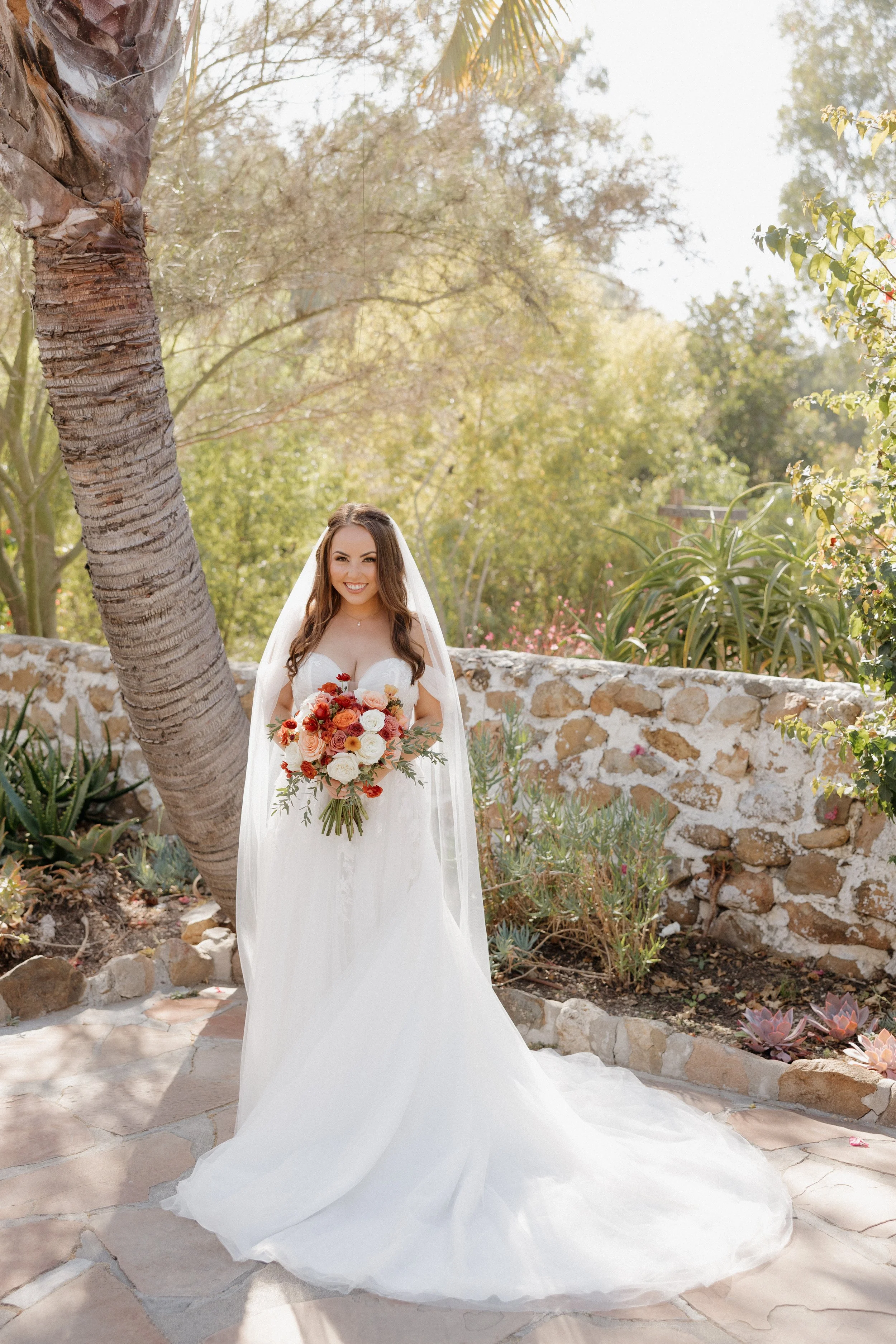 A bride in a white wedding dress and veil holding a vibrant bouquet of roses, standing outdoors near a stone wall and a large palm tree, surrounded by greenery and sunlight.