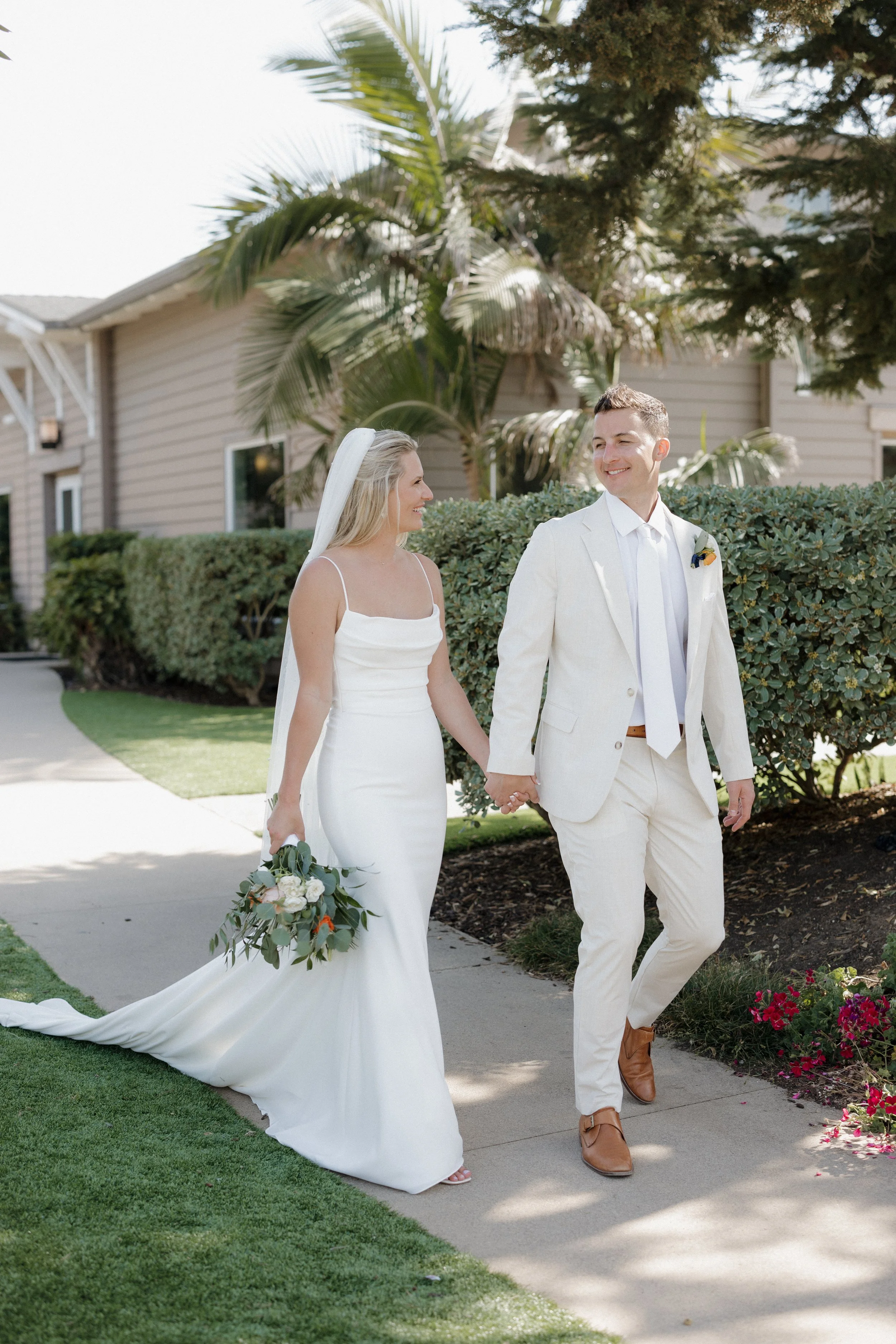 A bride and groom walking hand in hand outdoors on a sunny day, smiling at each other. The bride is wearing a white wedding dress and holding a bouquet, while the groom is dressed in a light-colored suit with a boutonniere. There are green plants and a house in the background.