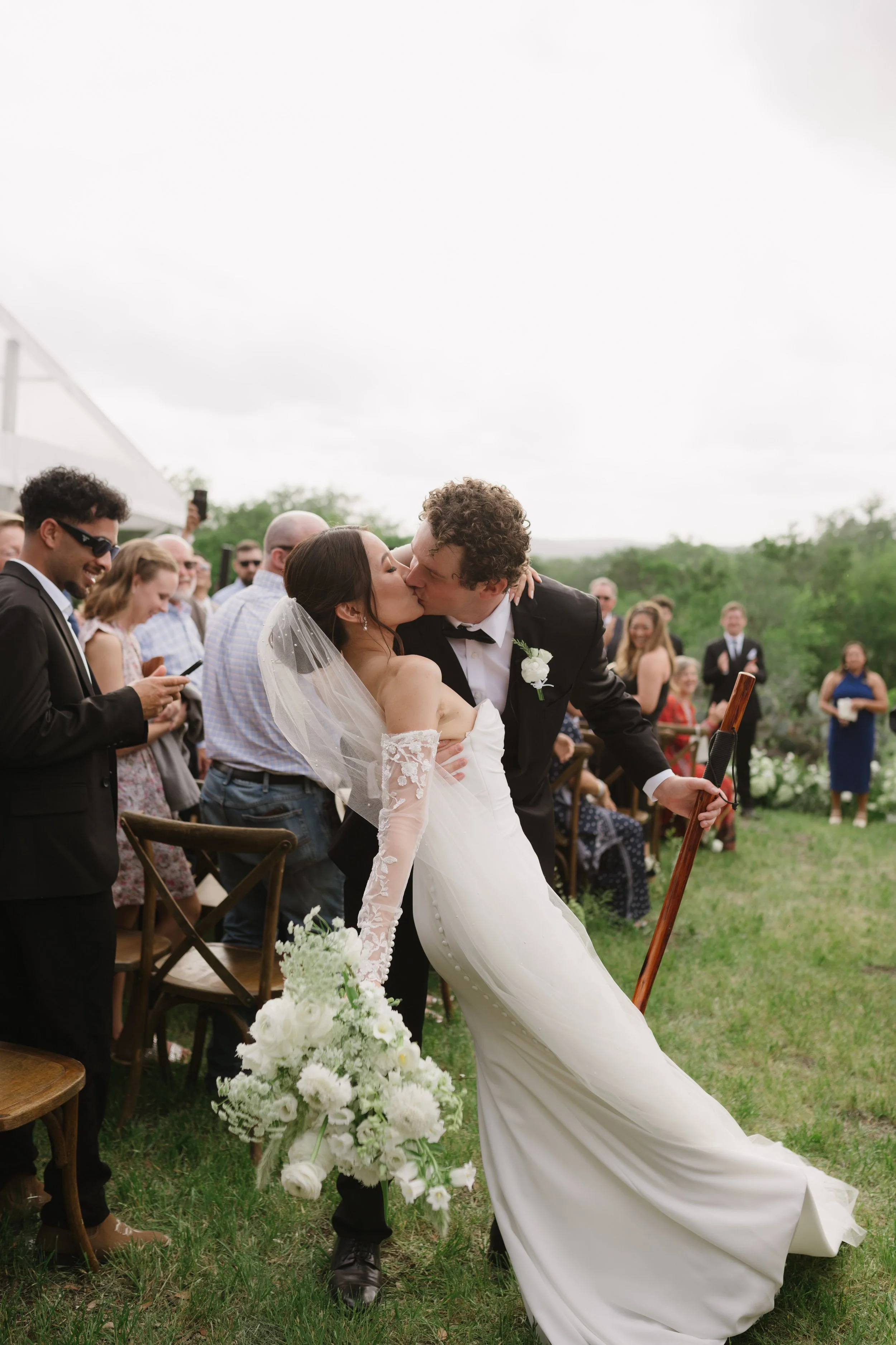 Bride and groom kissing outdoors at wedding ceremony, surrounded by guests.