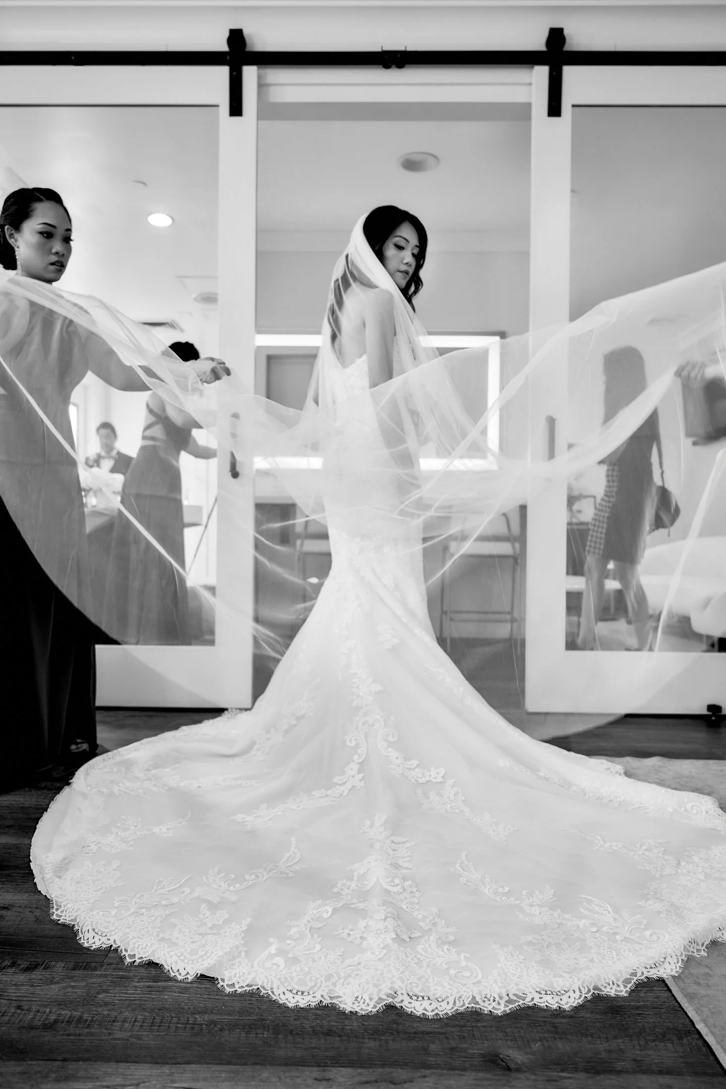 A bride in a wedding dress with a long train, surrounded by bridesmaids helping her with her veil, in a fitting room or bridal studio.