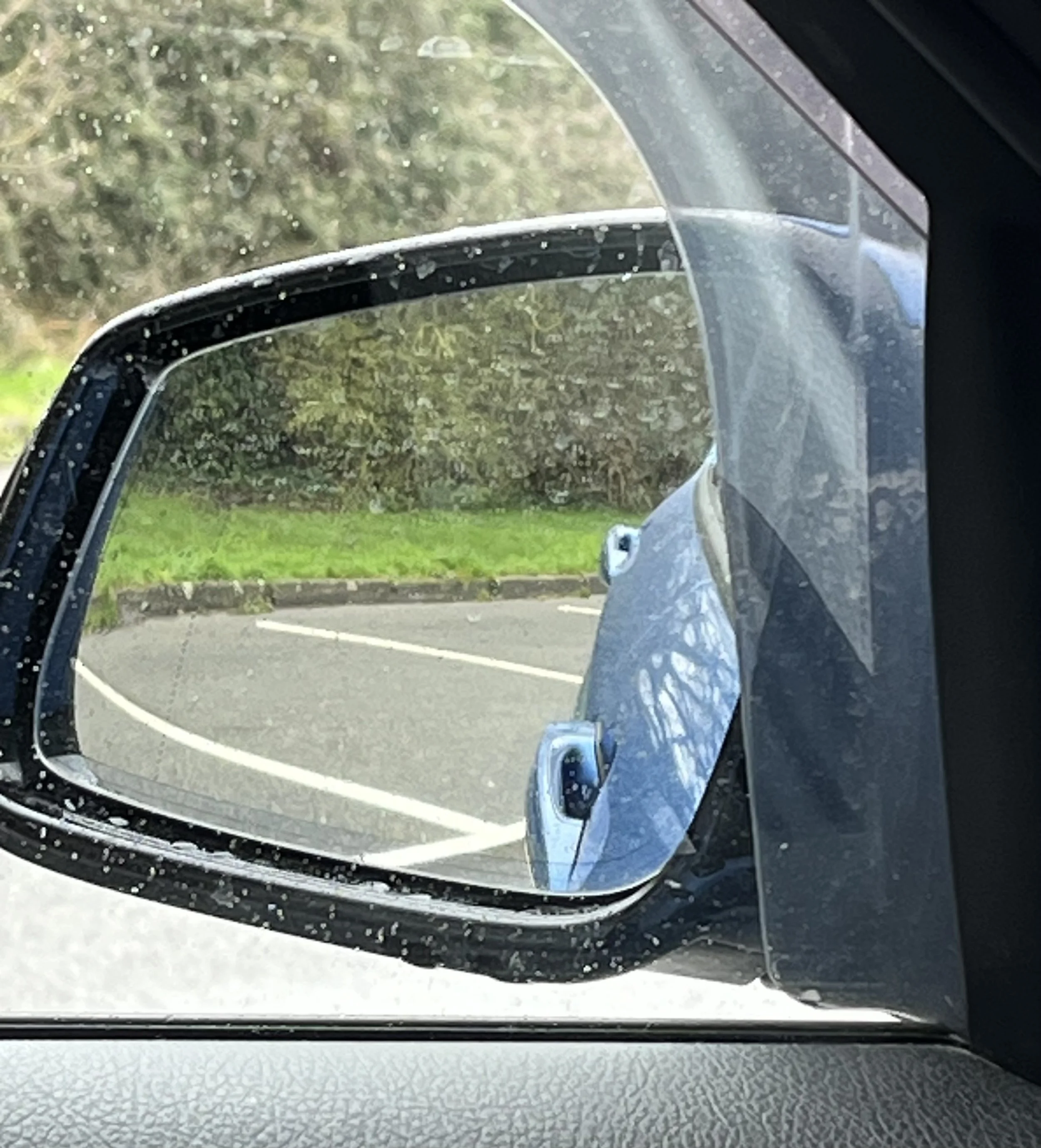 Car side mirror with a protective shield, showing a parking lot and greenery outside, with rain droplets on the mirror.