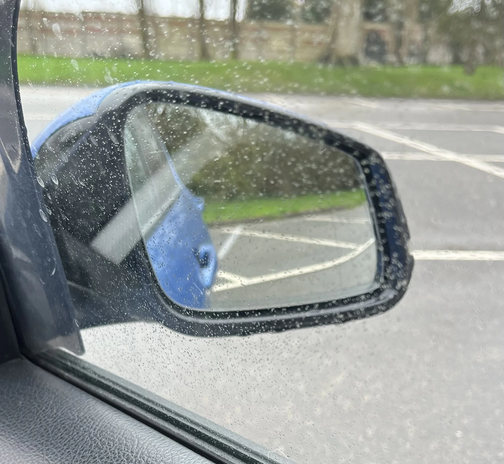 A car's side mirror with raindrops on it, showing a rainy parking lot with painted lines and a grassy area with trees in the background.