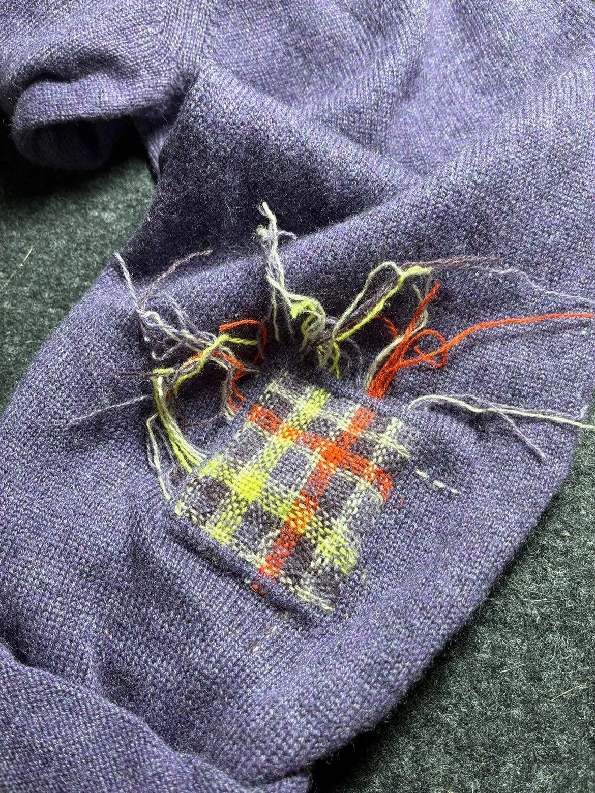 Ein beschädigtes, dunkelblaues Stoffstück mit losen, bunten Fäden und einem kleinen, abgenutzten karierten Patch.