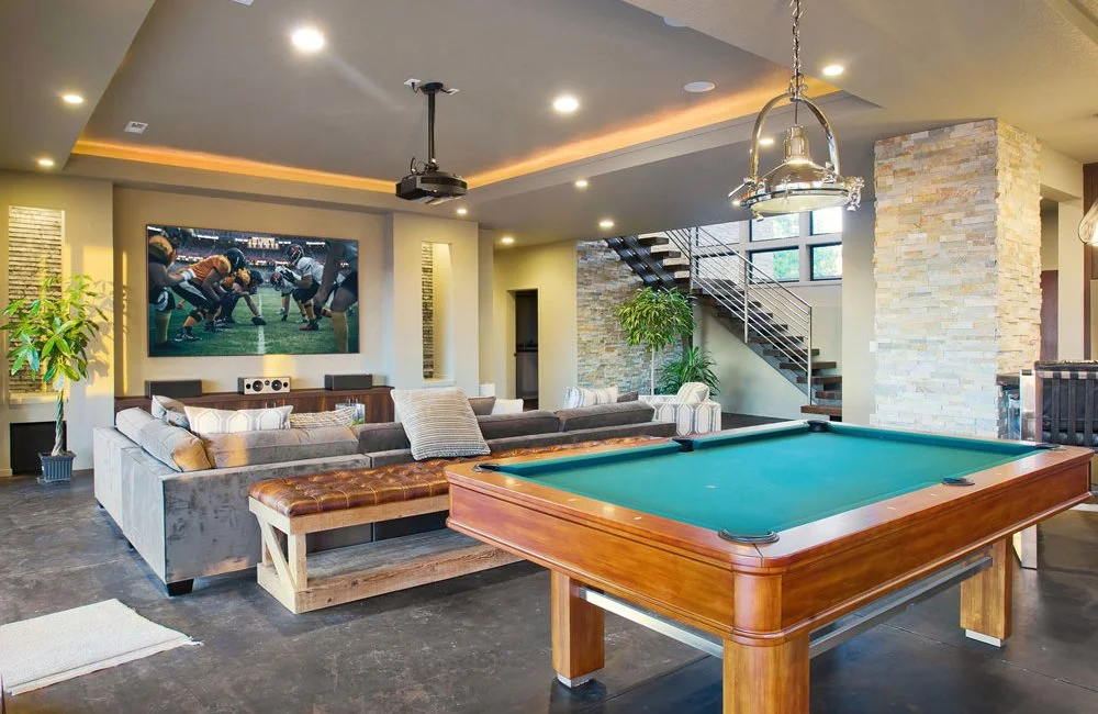 A modern living room with a pool table, gray sectional sofa, large TV showing a football game, and a staircase with a metal railing.