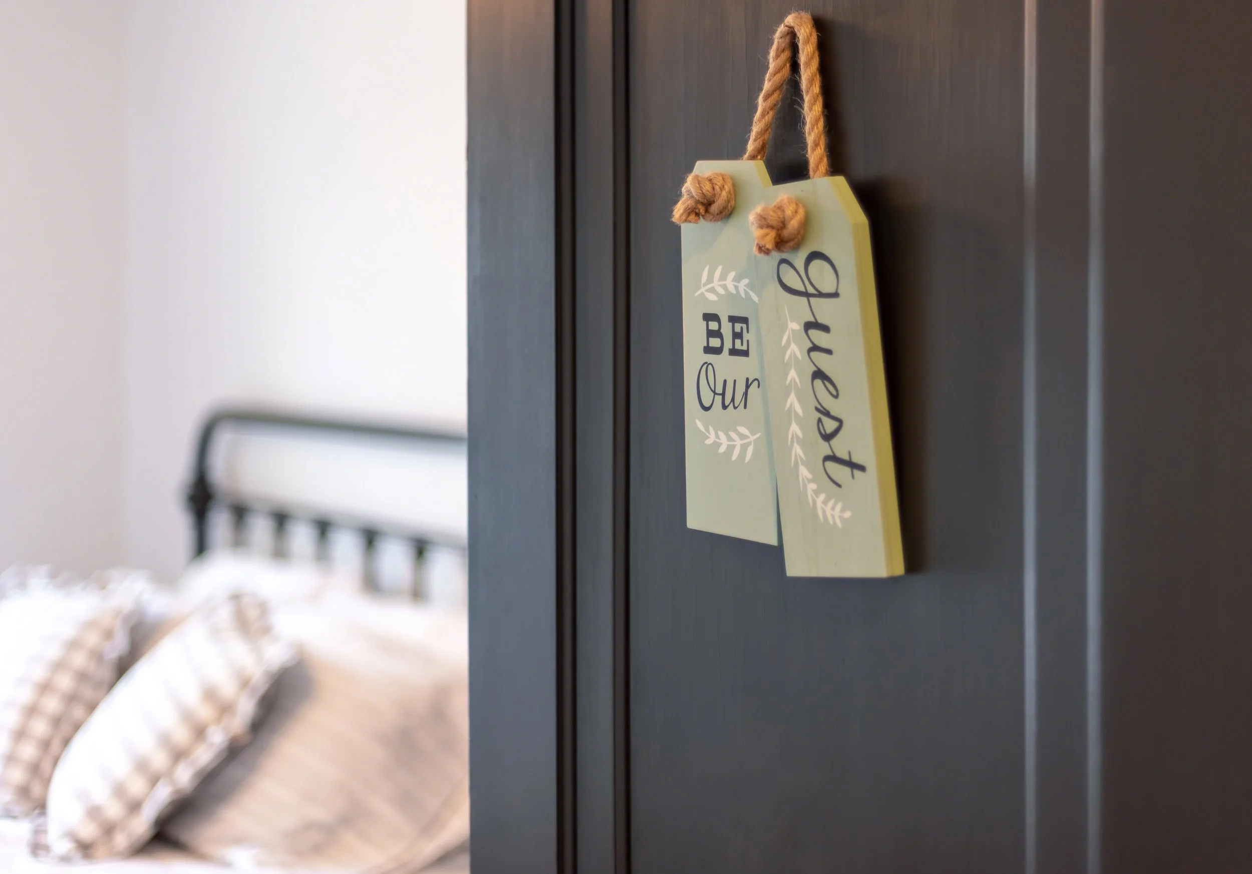 A black door with a decorative sign hanging from a rope that says "Be Our Bethened" in white and black text. In the background, a bed with a white headboard and beige pillows with checkered covers.