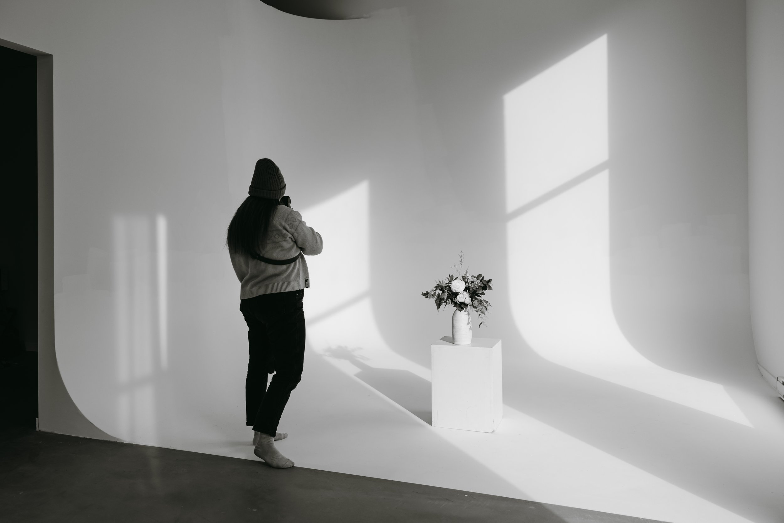 Une femme portant un bonnet et un pull blanche tient un téléphone, dans une pièce blanche avec une plante en vase sur un socle, et des jeux d'ombres et de lumières sur le mur.