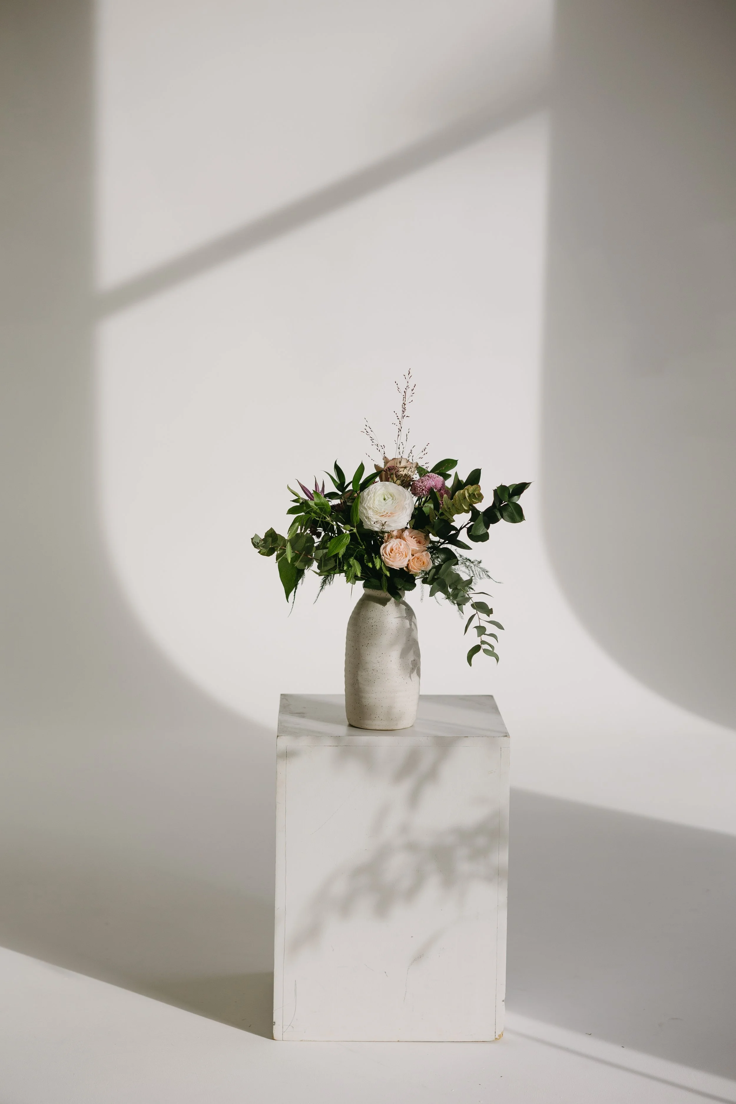 Vase en céramique blanche contenant un arrangement floral avec des roses, des feuillages verts et des fleurs de couleur pastel, placé sur un socle blanc.