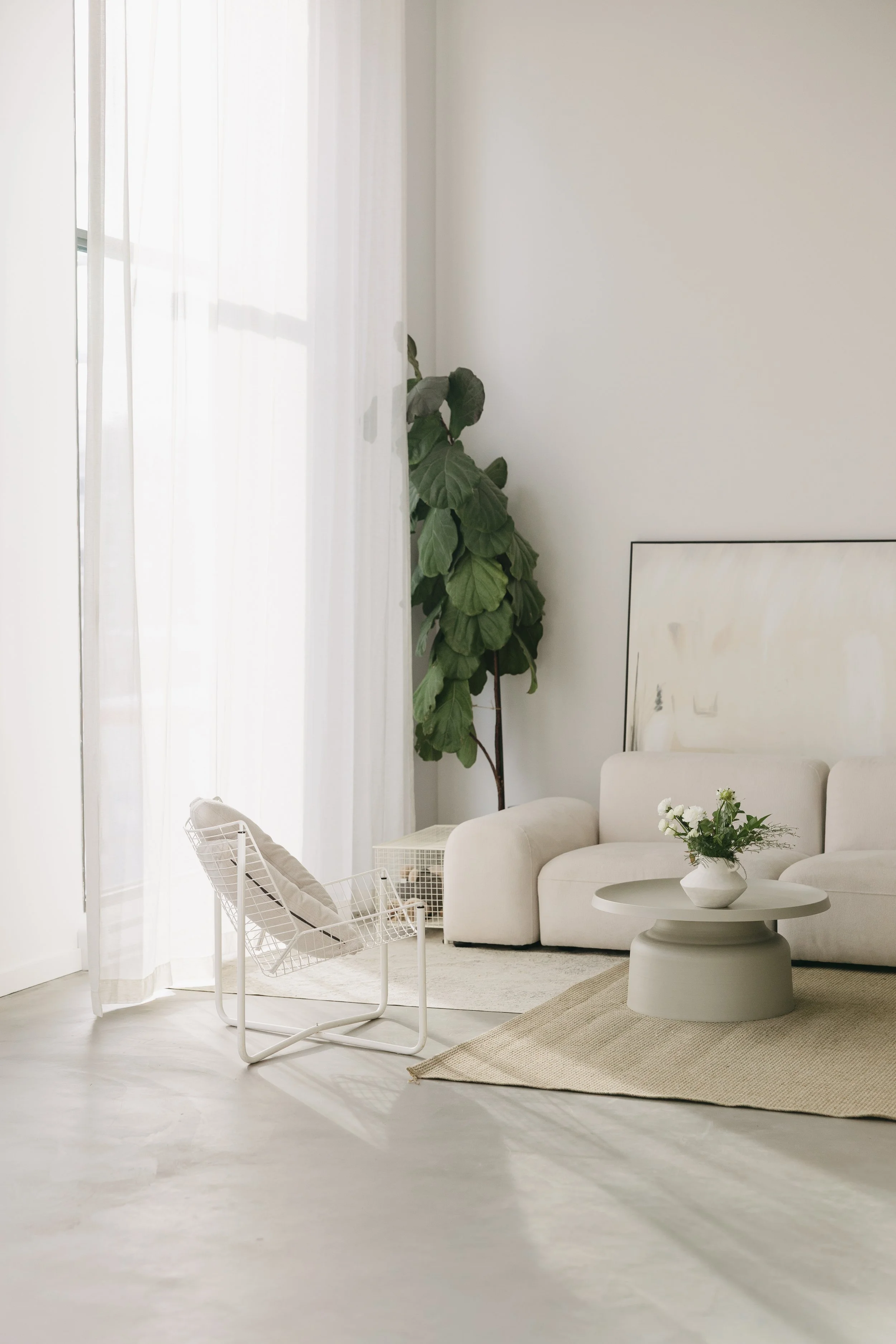 Salon minimaliste avec canapé blanc, table basse ronde, vase de fleurs blanches, grande plante verte dans un coin, et rideaux blancs laissant passer la lumière