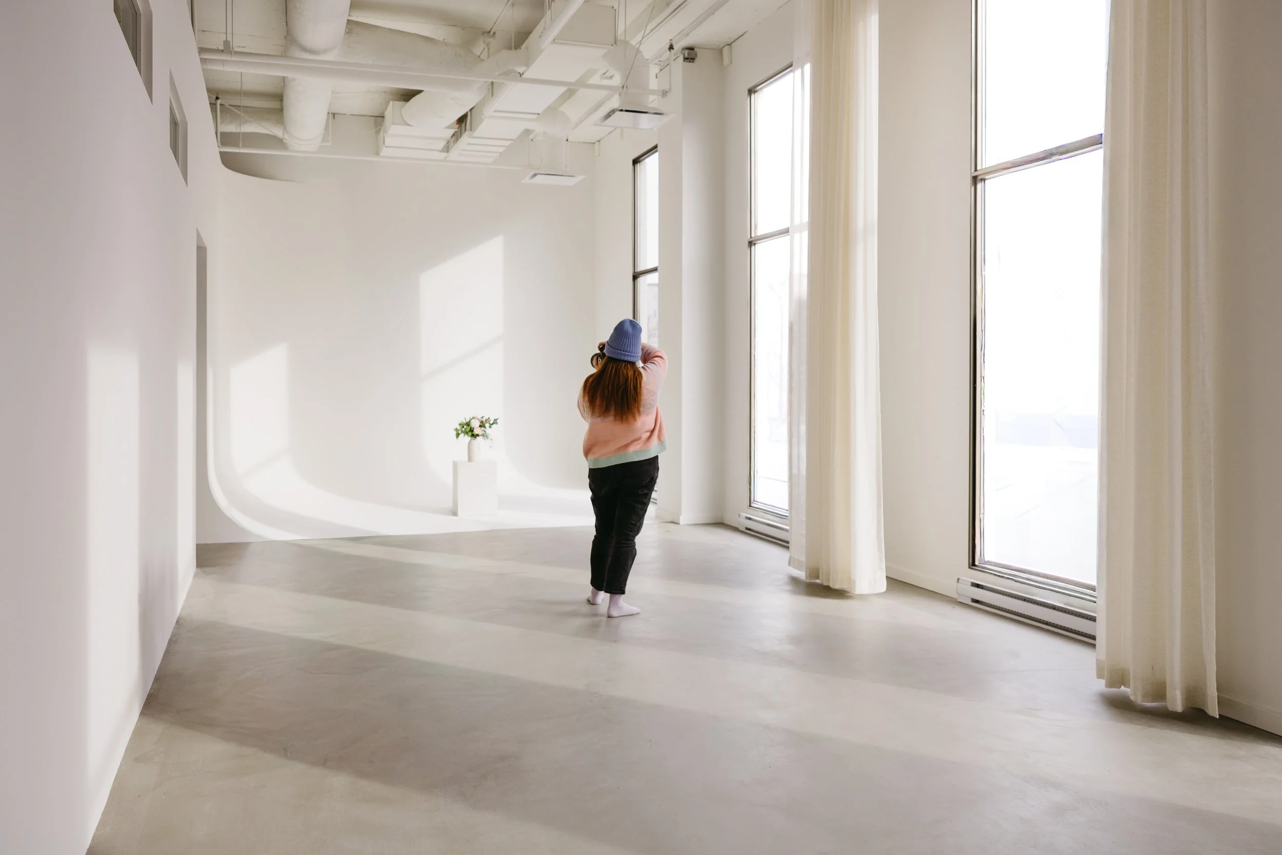 Une femme portant un chapeau bleu, un pull rose et un pantalon noir se tient dans une pièce lumineuse avec de grands fenêtres, des rideaux blancs et un vase avec des fleurs sur un piédestal blanc.