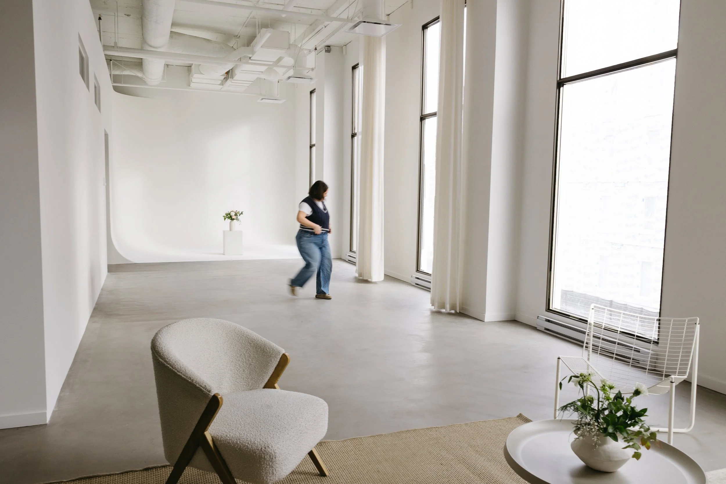 Une femme marche dans une pièce lumineuse avec de grands fenêtres, un fauteuil beige, une table avec une plante, et une chaise blanche.