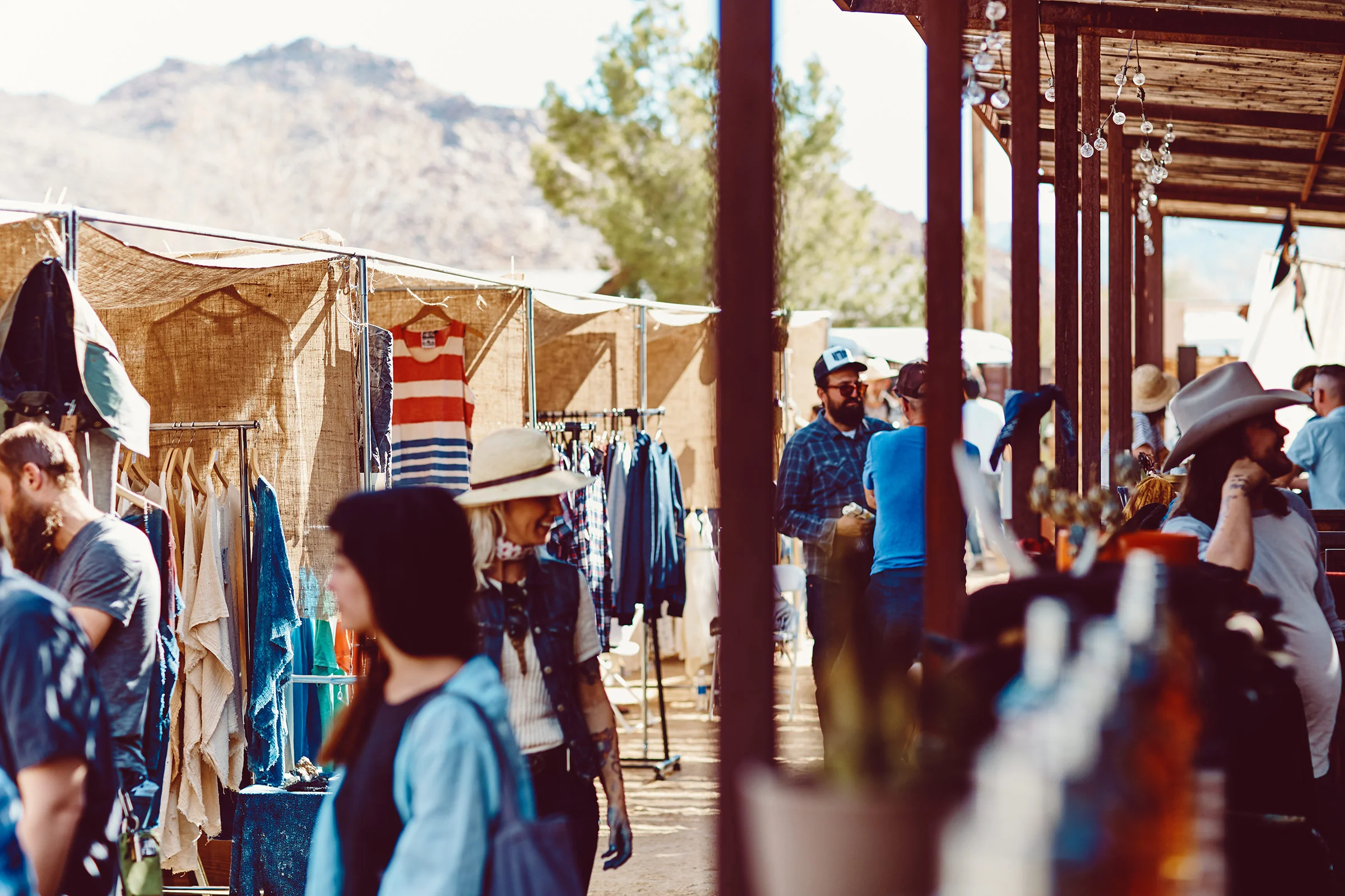 Mercadillo al aire libre con puestos de ropa y personas paseando y comprando, en un entorno montañoso y soleado.