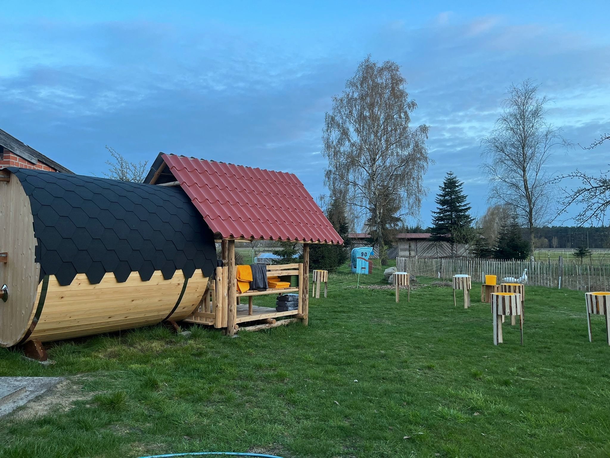 Garten mit einer Sauna in Fassform mit schwarzem Dach und einem überdachten Sitzbereich mit rotem Dach. Mehrere runde Holztische auf einer grünen Wiese. Im Hintergrund ein Holzzaun und Bäume.