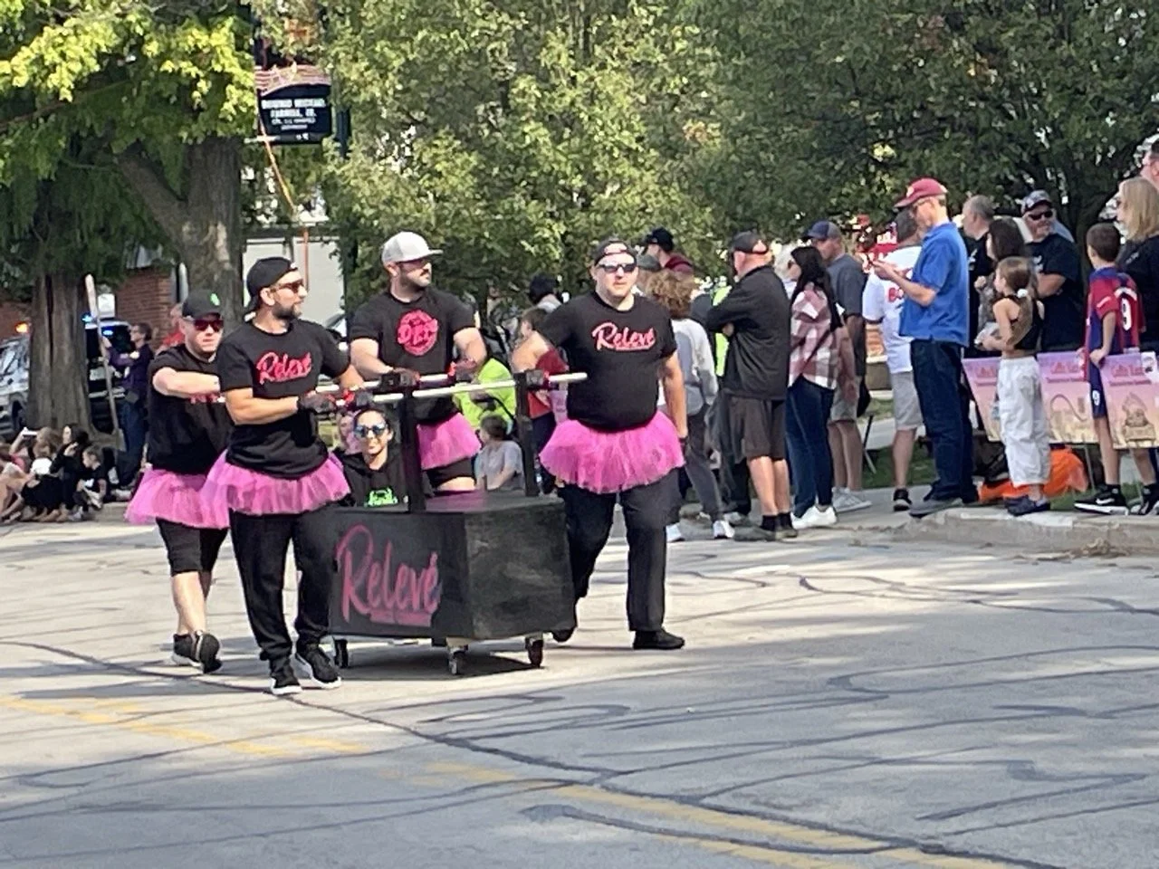 Group of Relevé Dance Company performers representing Lockport and surrounding areas like Joliet and Romeoville, participating in a parade dressed in black shirts and pink tutus.