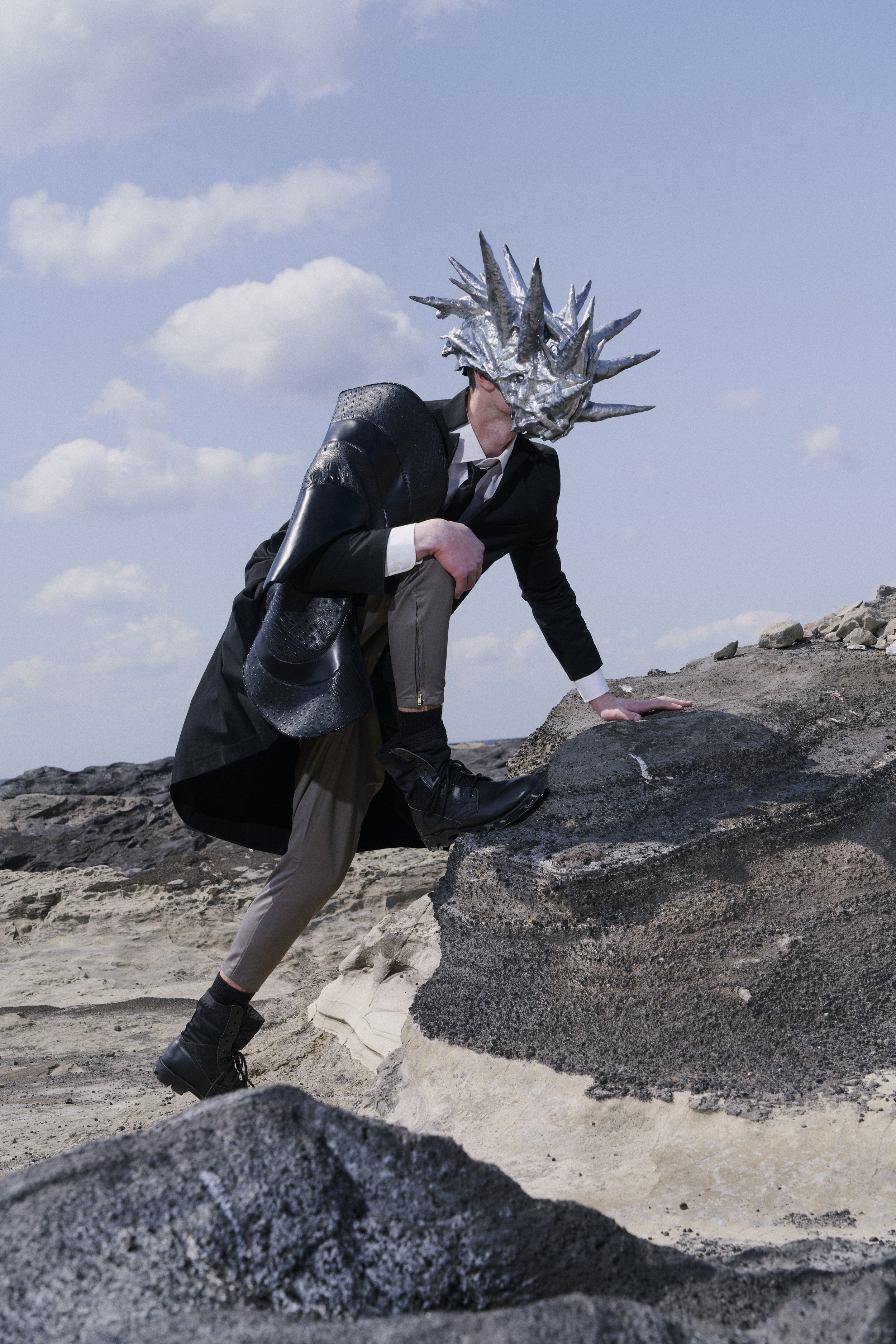 Person wearing a suit and a metallic spiked dragon mask, climbing a large rock on a rocky terrain with a cloudy sky background.