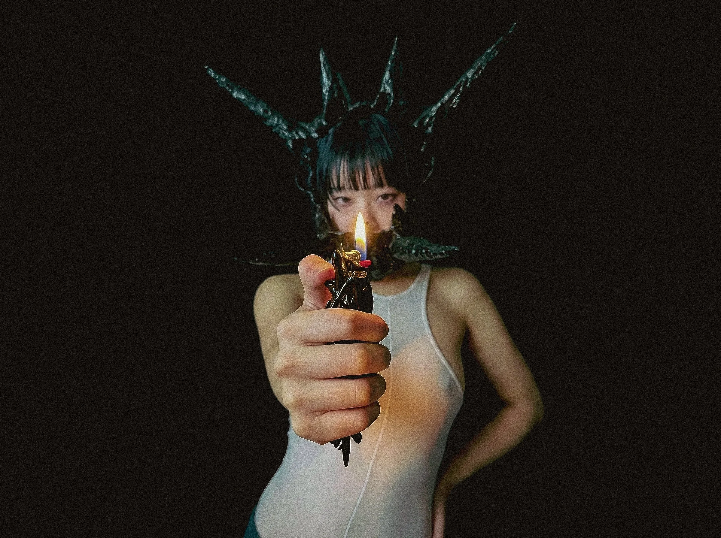 A person with black hair and bangs, wearing a white sleeveless top, holds a lighter with a small flame facing the camera, against a black background.