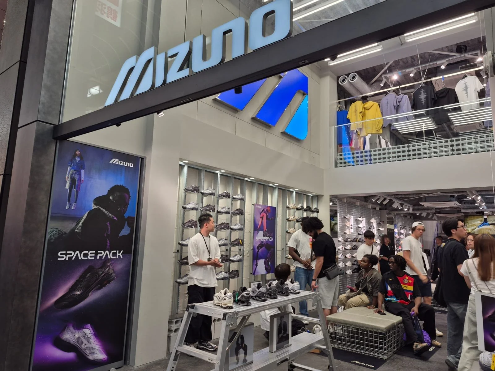 Inside a Mizuno store with clothing on the upper level, shoes on display below, and customers shopping or waiting in line.