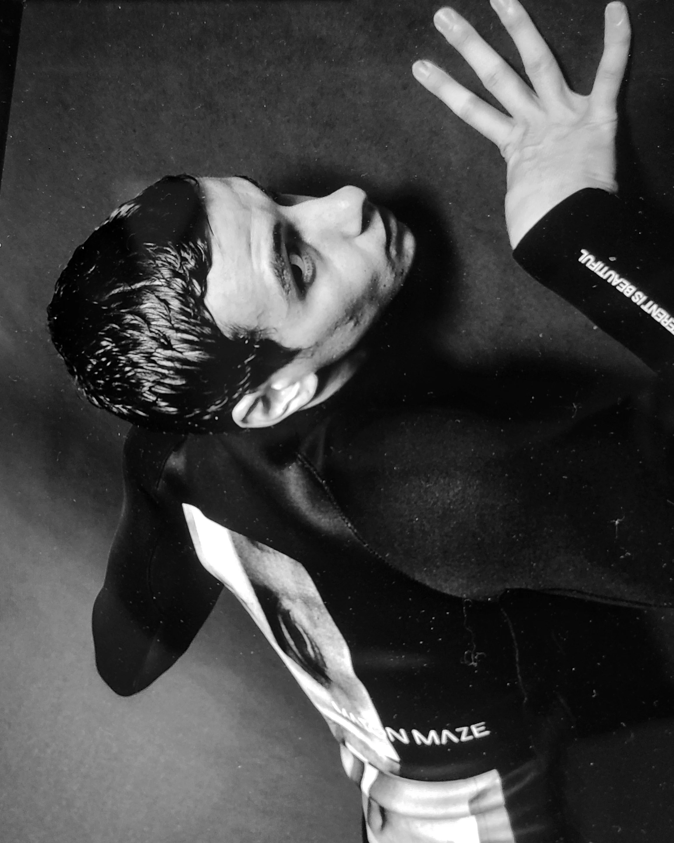 A man with wet, dark hair lying on his side on a dark surface, wearing a sports jacket with the words 'SPORTS BEAUTIFUL' on the sleeve. His left hand is extended outward, palm down, and he is looking upward at the camera.