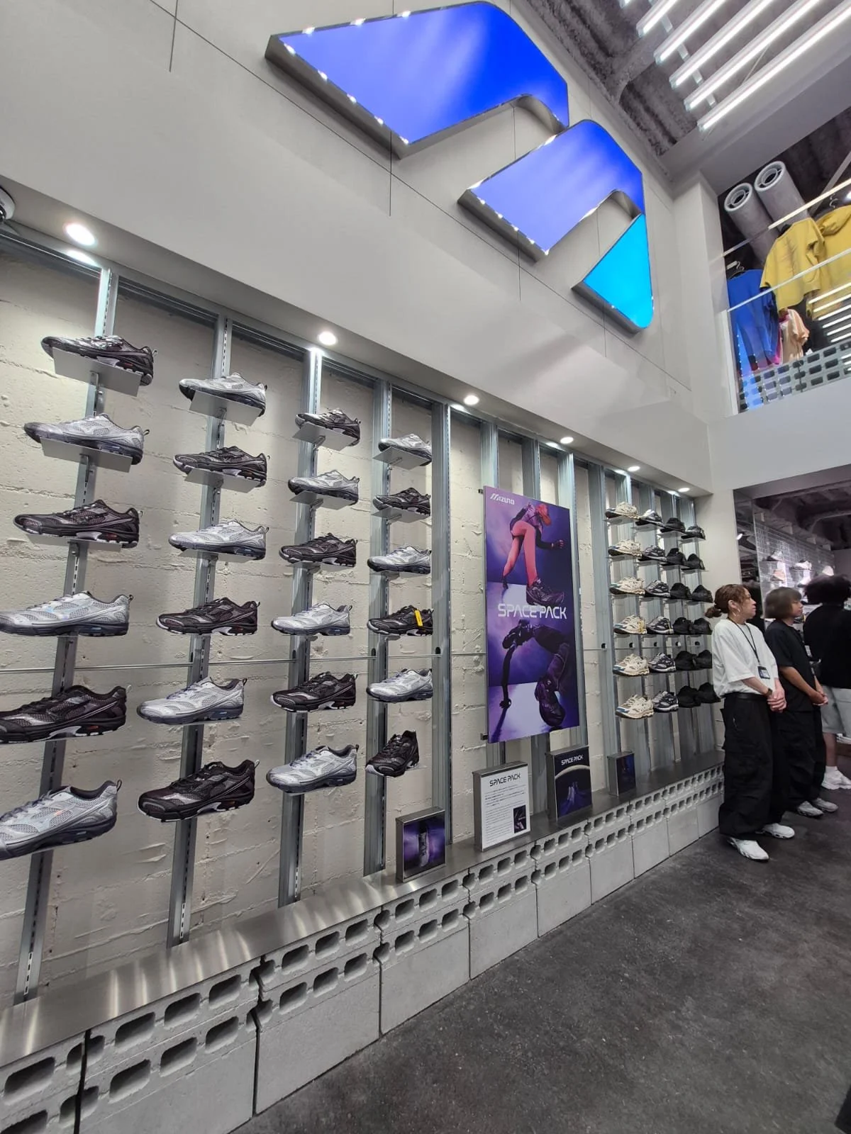A retail store display featuring multiple rows of men's athletic shoes on metal racks, a promotional poster for 'Space Pack' shoes, and a few customers standing nearby.