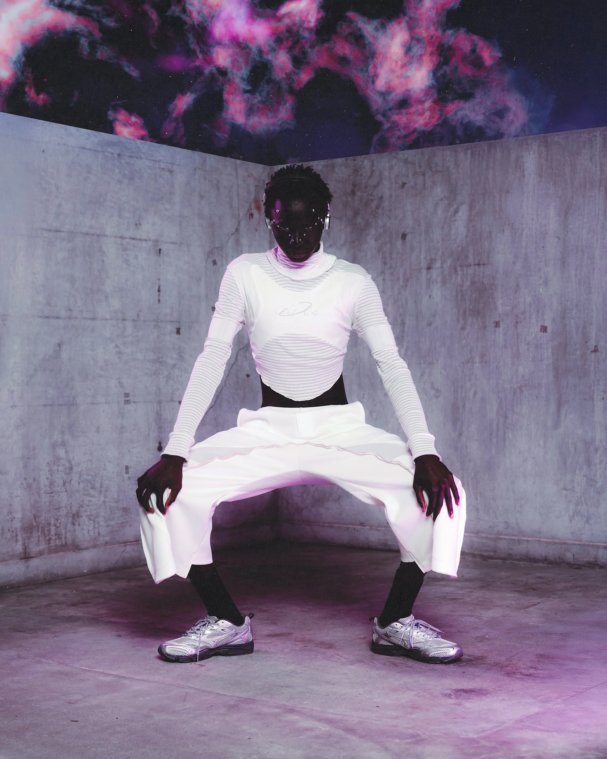 A person with dark skin and dreadlocks wearing a futuristic white outfit with metallic sneakers, squatting in a concrete room with a colorful nebula and stars projected on the ceiling.