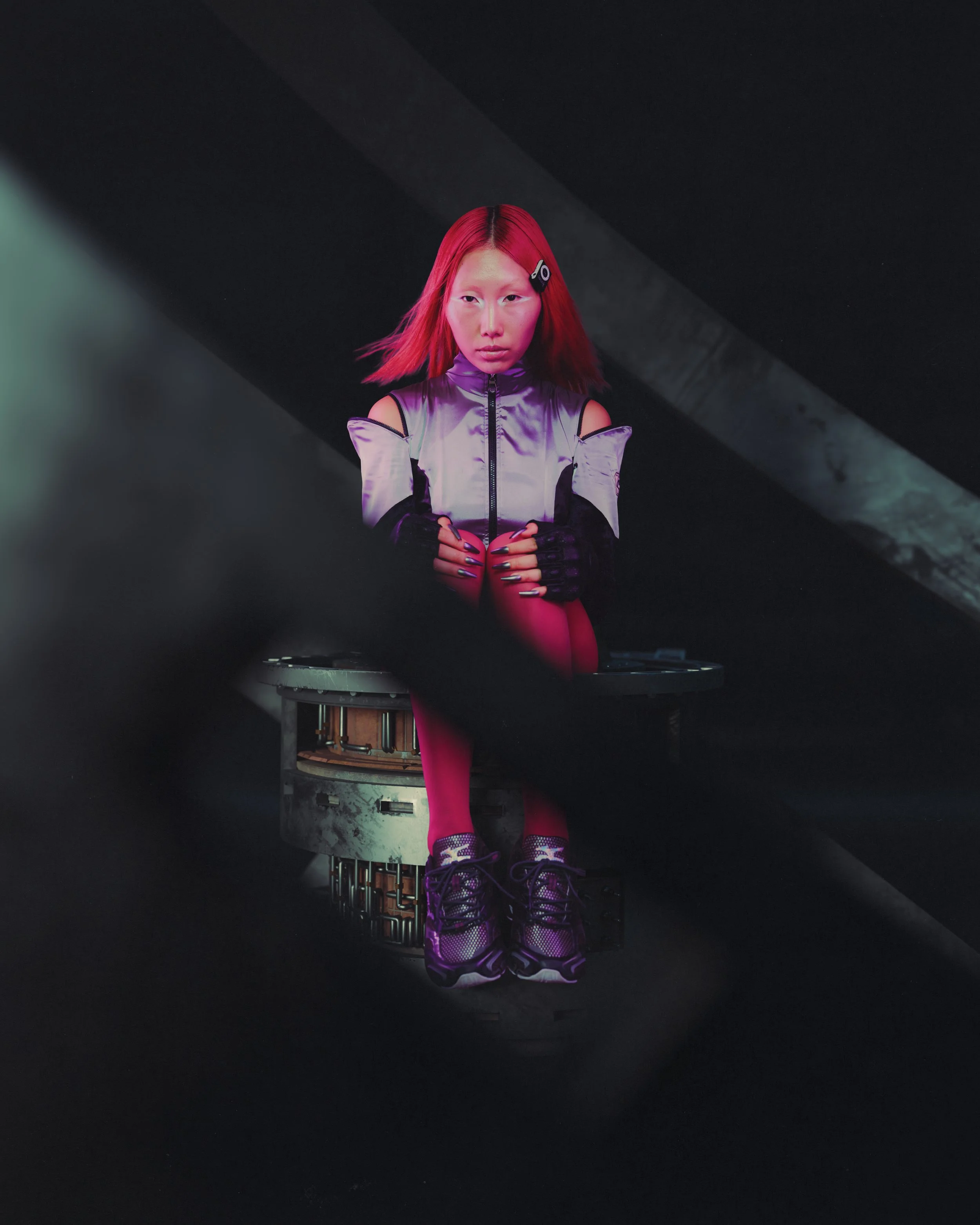 Young woman with red hair sitting on a mechanical platform against a dark background, wearing a shiny silver and black jacket, pink tights, black gloves, and purple running shoes.