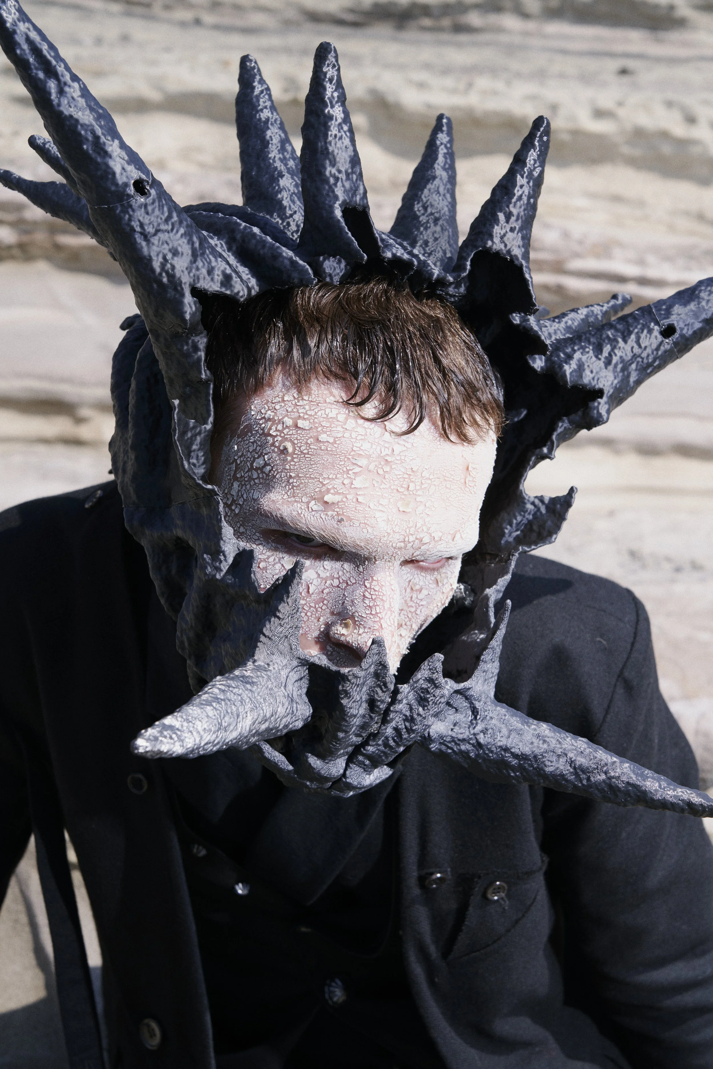 Person with cracked white face paint wearing a spiked black headpiece and gloves, sitting outdoors on a sandy surface.