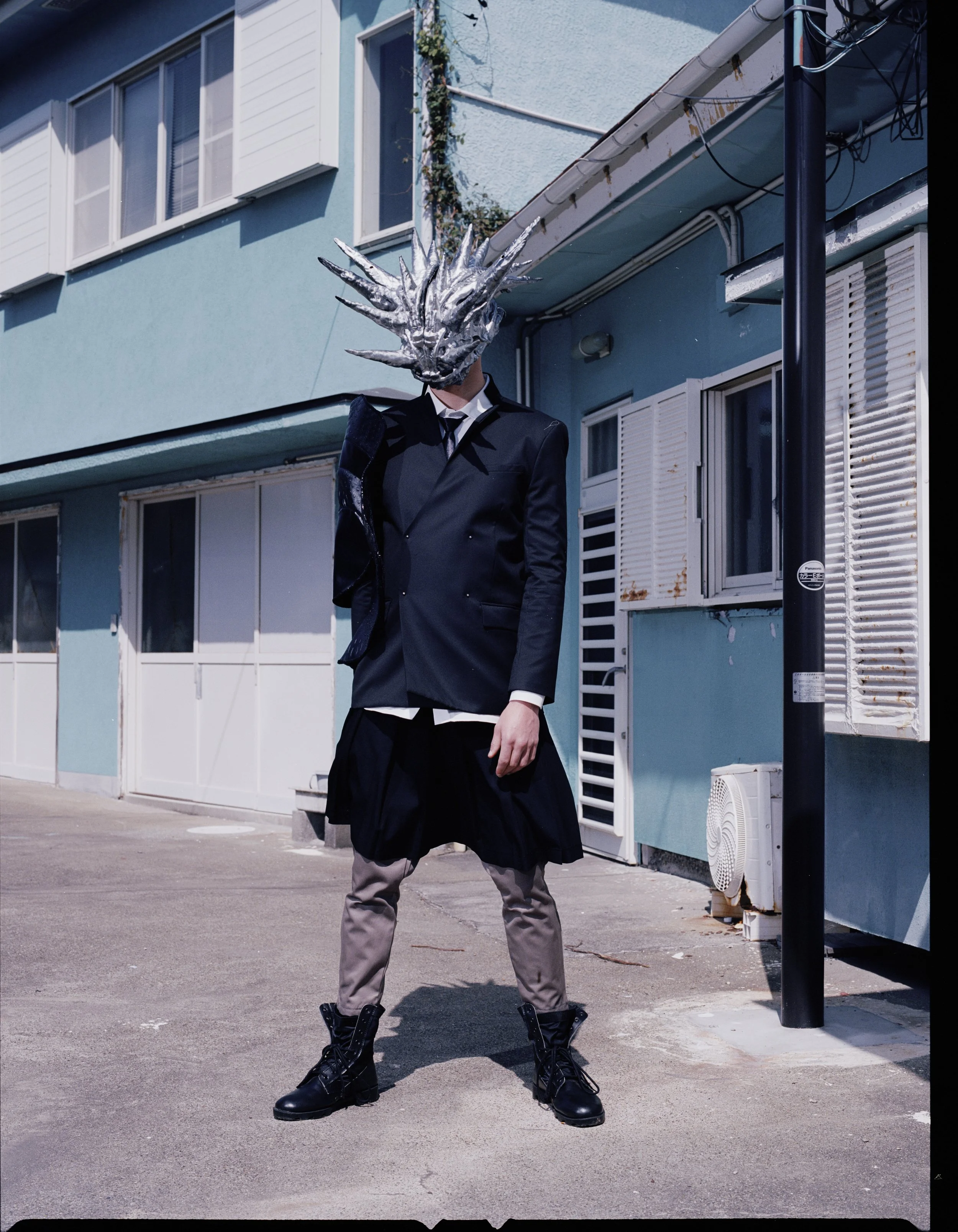 Person in a black suit with a large, ornate silver dragon mask standing on an urban sidewalk in front of a blue building with windows.