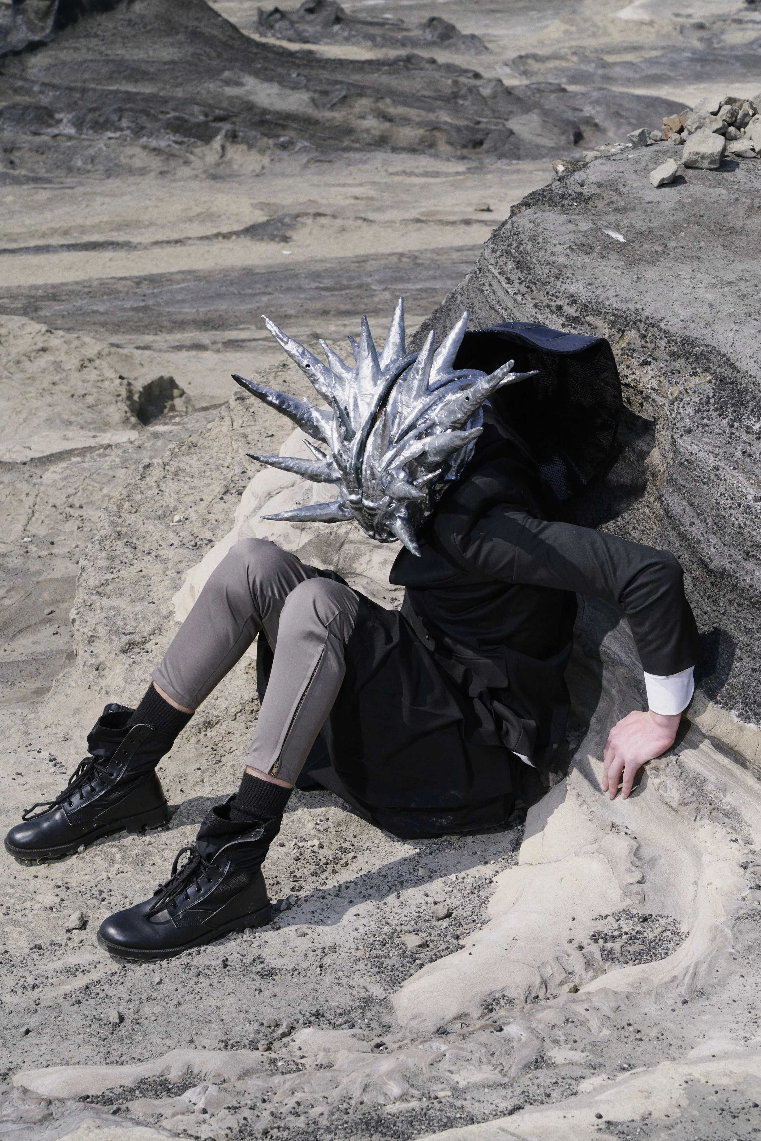 Person dressed in black with a head covered by a spiked, metallic silver mask, sitting on sandy terrain with rocks and barren landscape in the background.