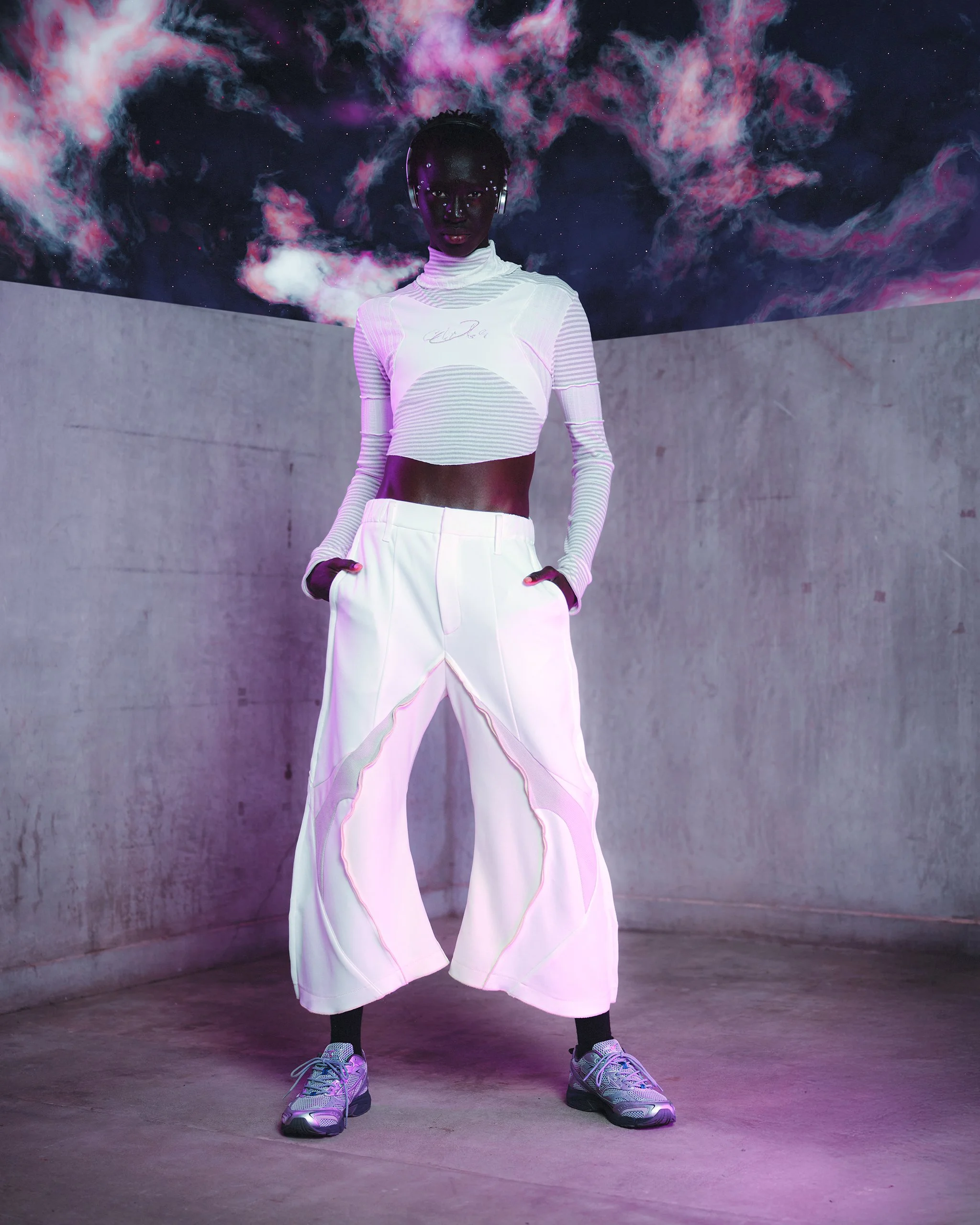 A woman with dark skin and short hair wearing a futuristic white crop top with long sleeves and wide-legged pants, standing in a minimalist space with a concrete wall and a galaxy-themed ceiling.