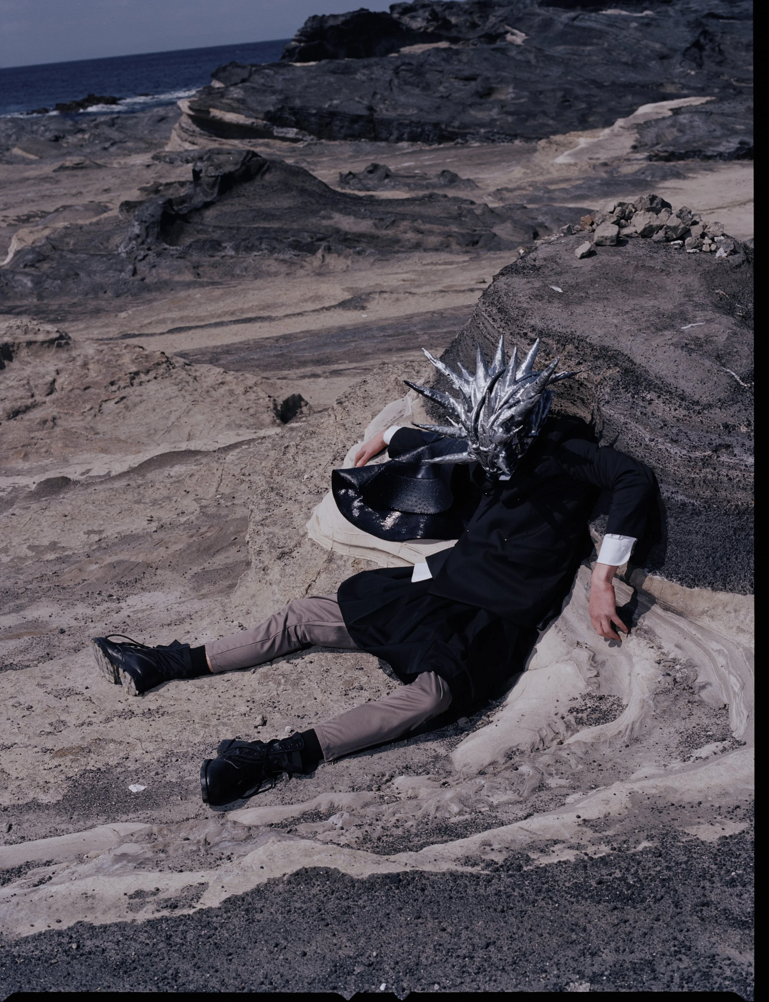 Person wearing a costume with a metallic dragon mask, black coat, and beige pants, sitting on rocky terrain near an ocean with a dark sky in the background.