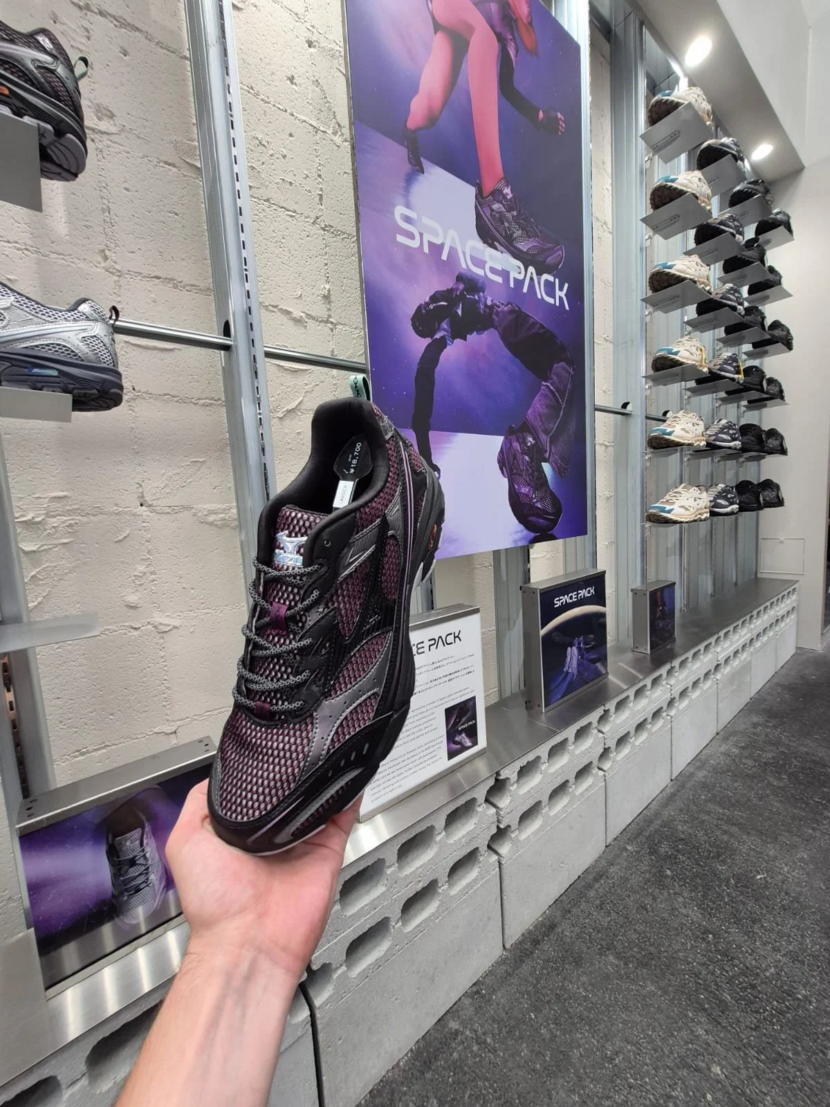 Person holding a futuristic sneaker in a store, with shelves of similar shoes and a large poster displaying the same shoe design.